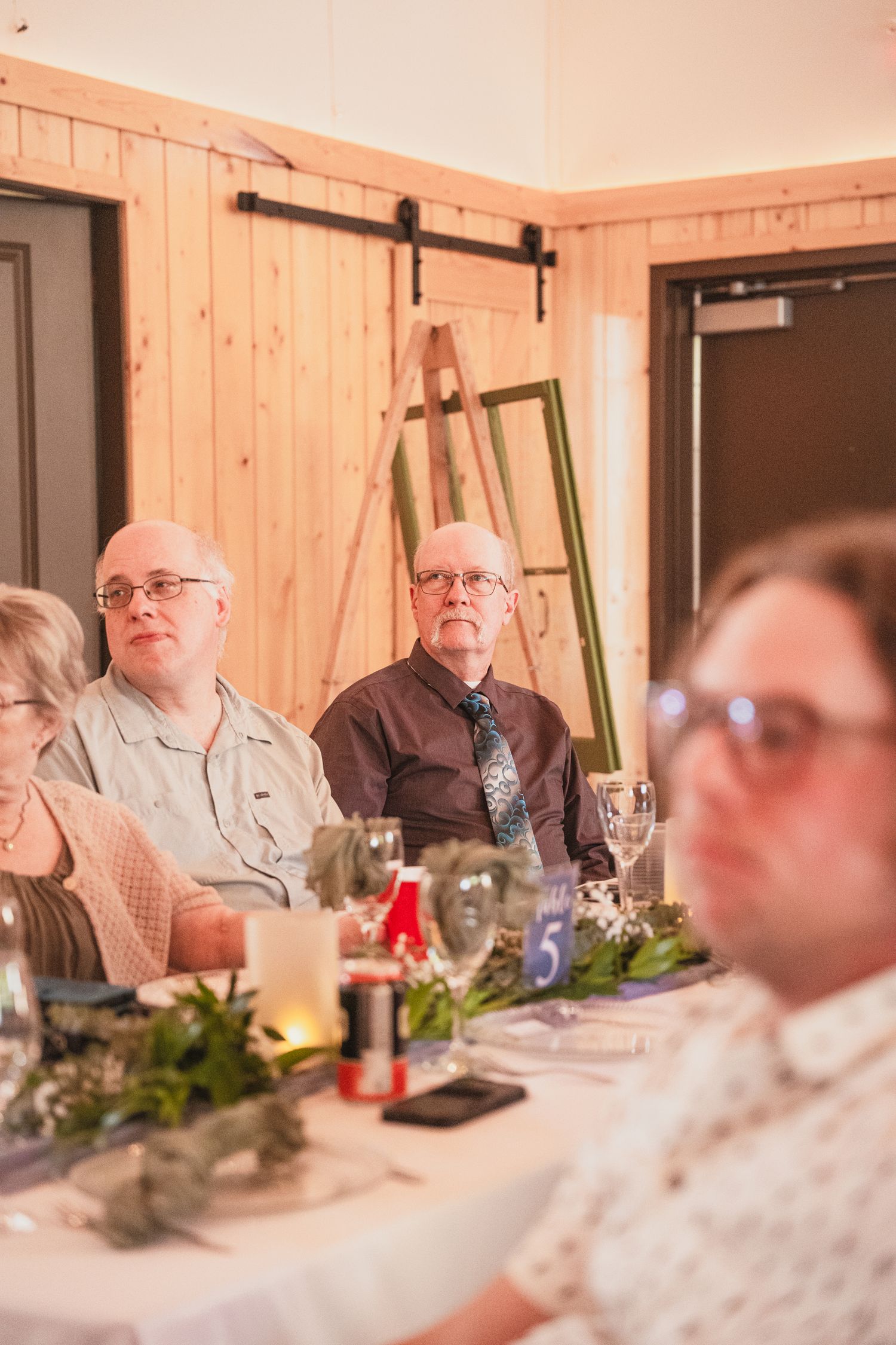 Wedding guests seated at a decorated reception table with greenery centerpieces in a rustic wood-paneled venue.