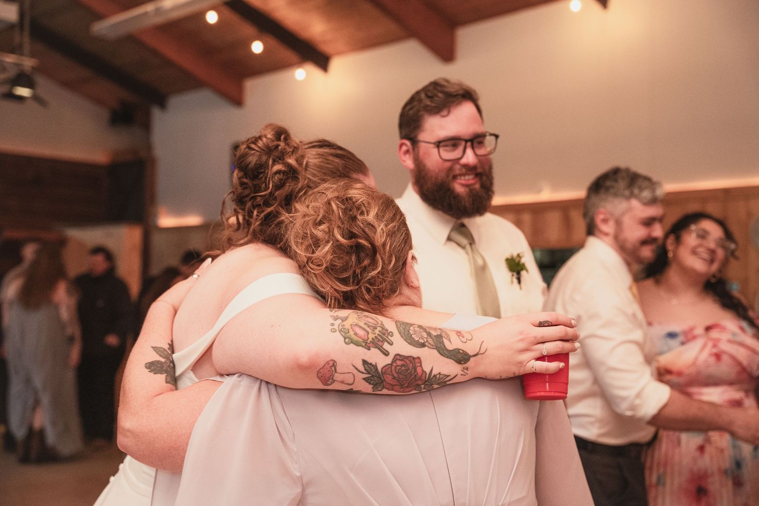 Bride hugging bridesmaid with tattoos at wedding reception as bearded groom smiles nearby in rustic venue.