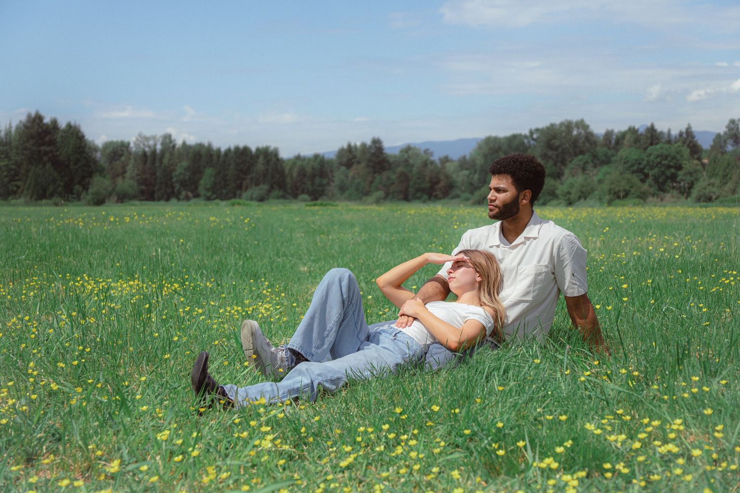 Couple relaxing in a sunny green meadow with wildflowers, man sitting while woman lies in his lap, trees in background.