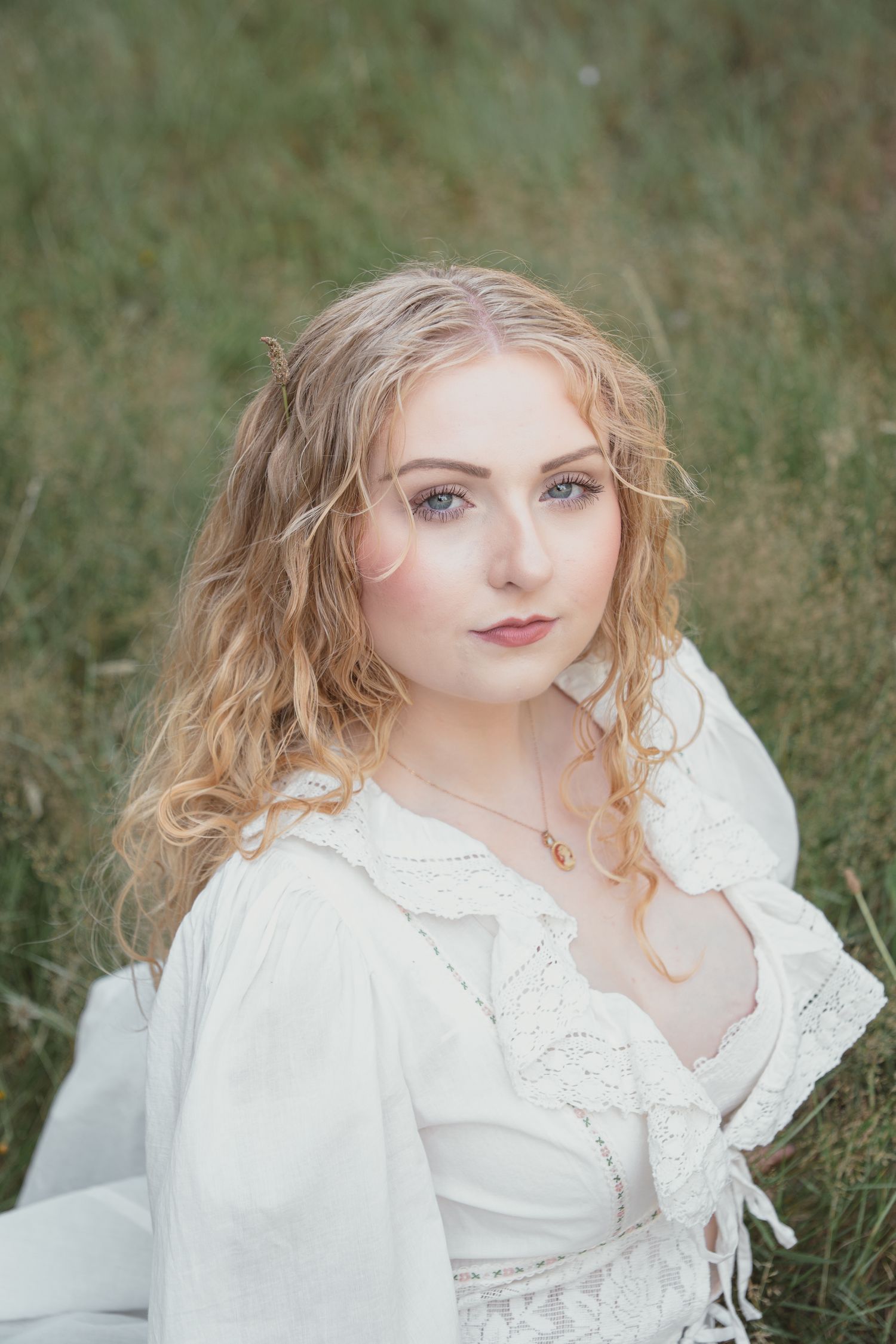 Young woman with curly blonde hair in white lace dress sitting in green field, wearing gold necklace.
