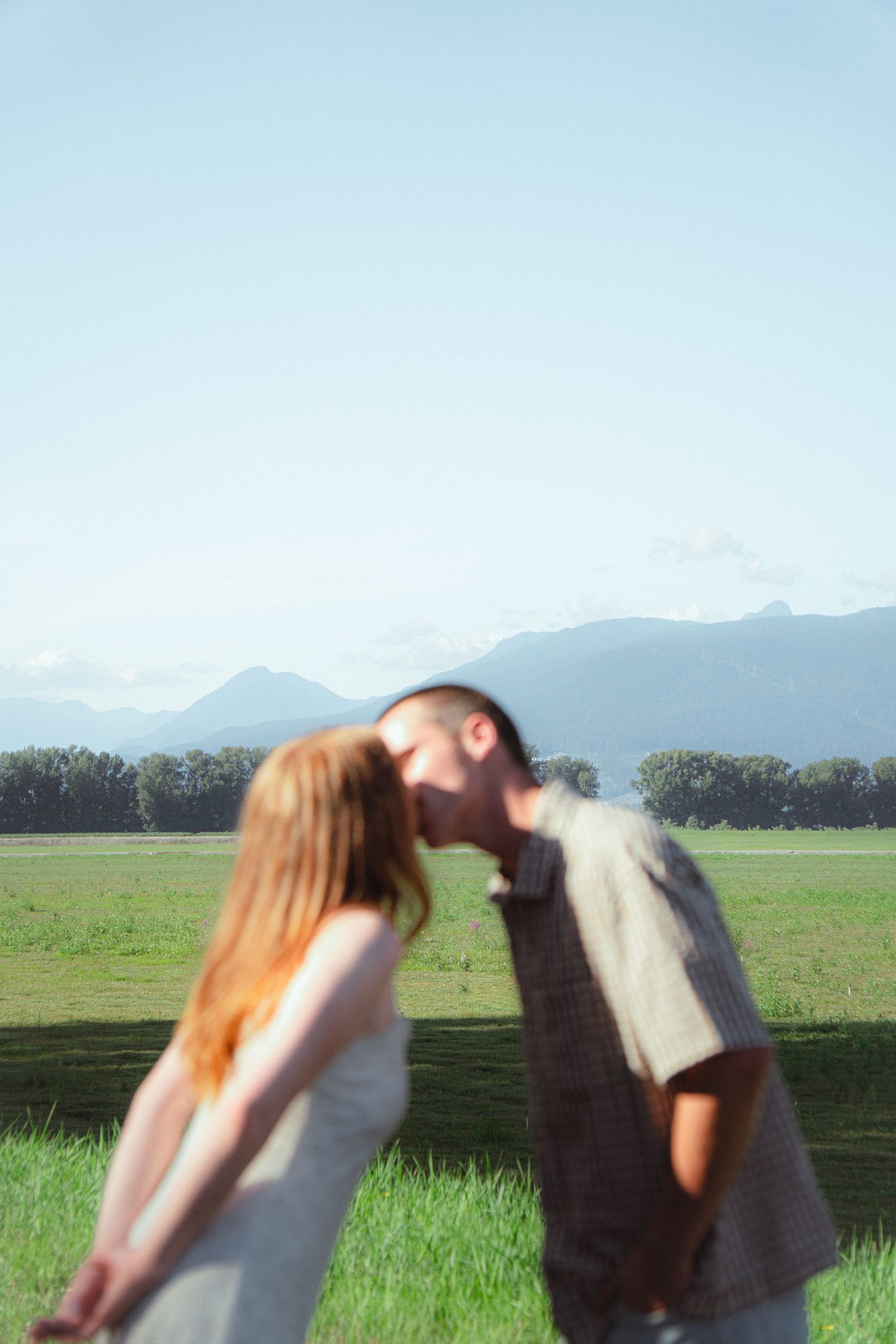 A couple shares a romantic kiss in a lush green field with mountains and clear sky in the background.
