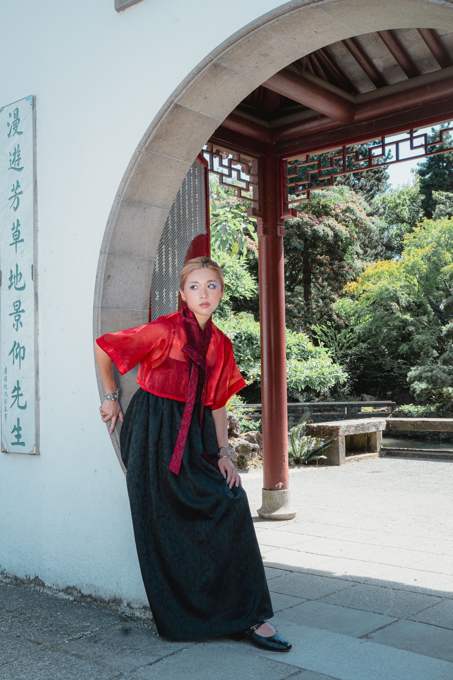 Woman in red and black traditional Chinese outfit stands in garden moon gate.