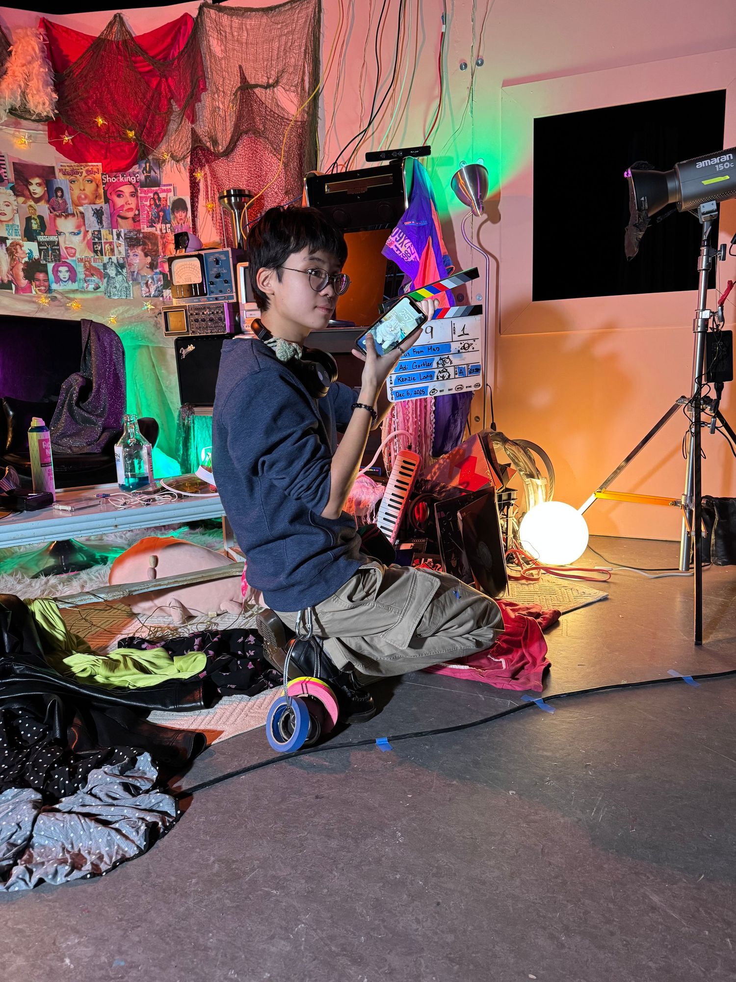 Film student kneels on colorful cluttered movie set holding props under vibrant neon lights.