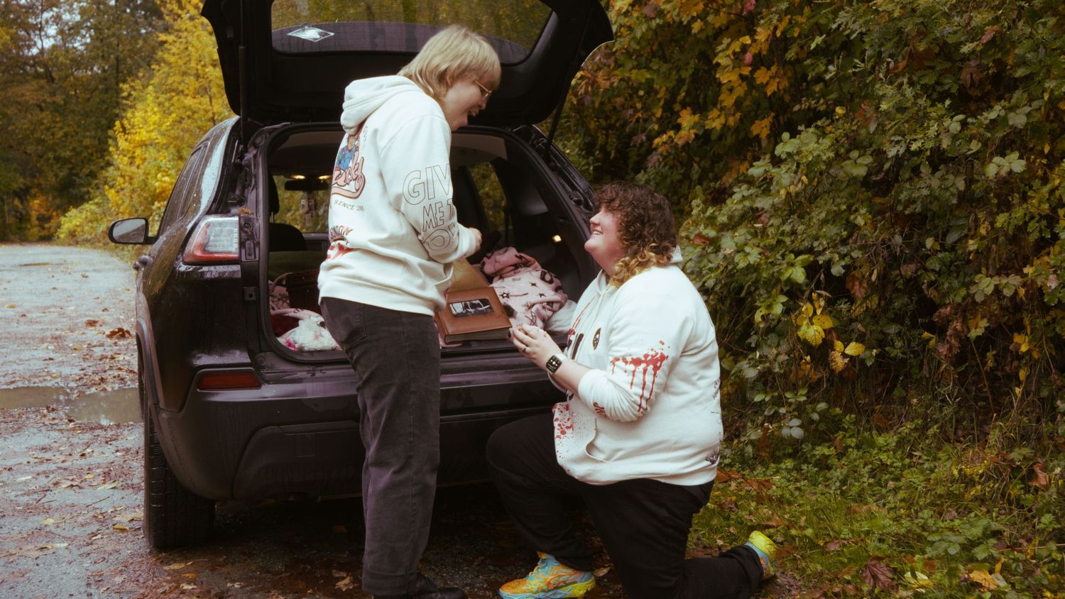 Two people in white graphic hoodies unload items from the trunk of a dark SUV on an autumn road.