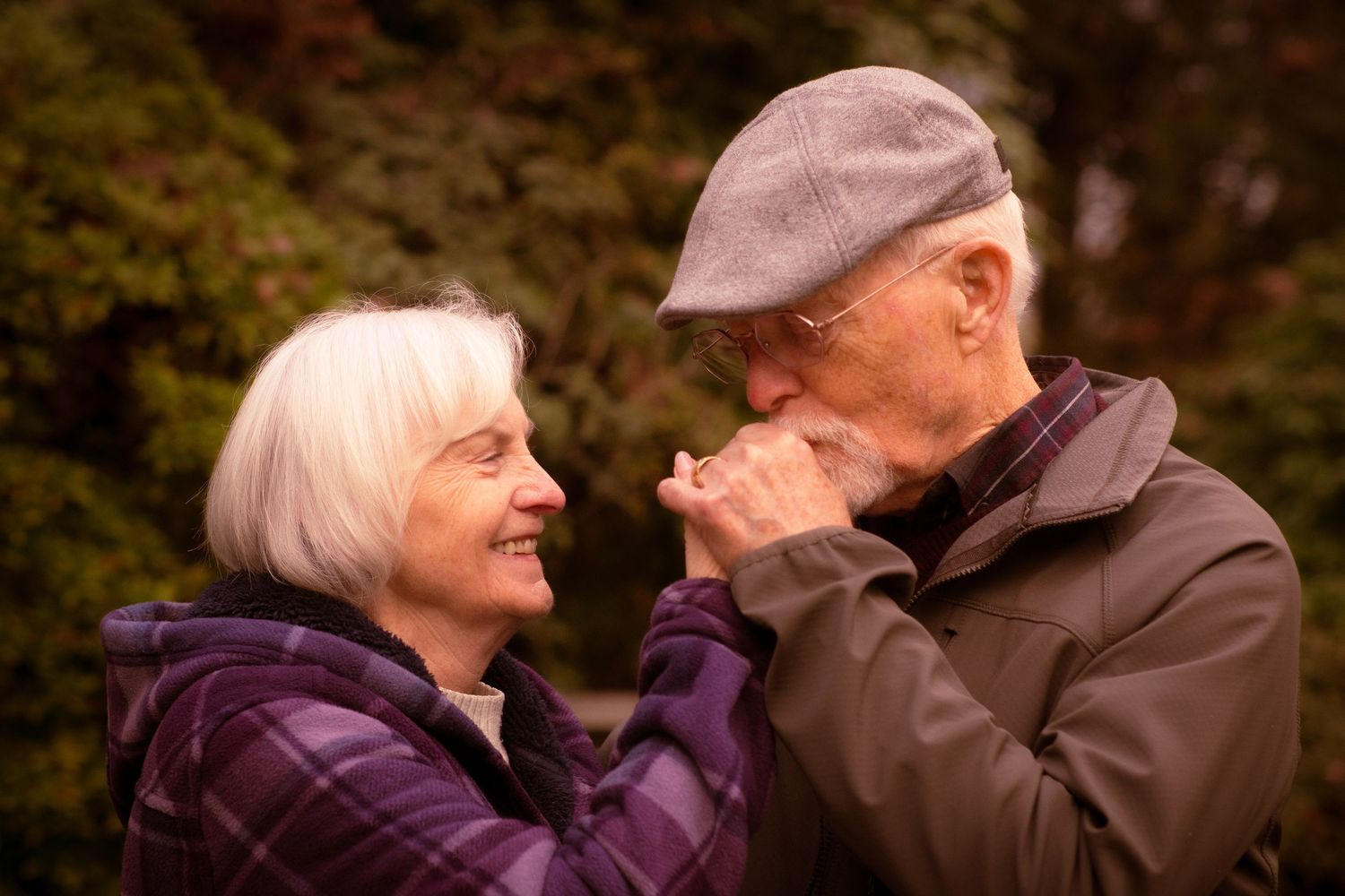 Senior man kissing woman's hand tenderly while she smiles in an outdoor autumn setting.