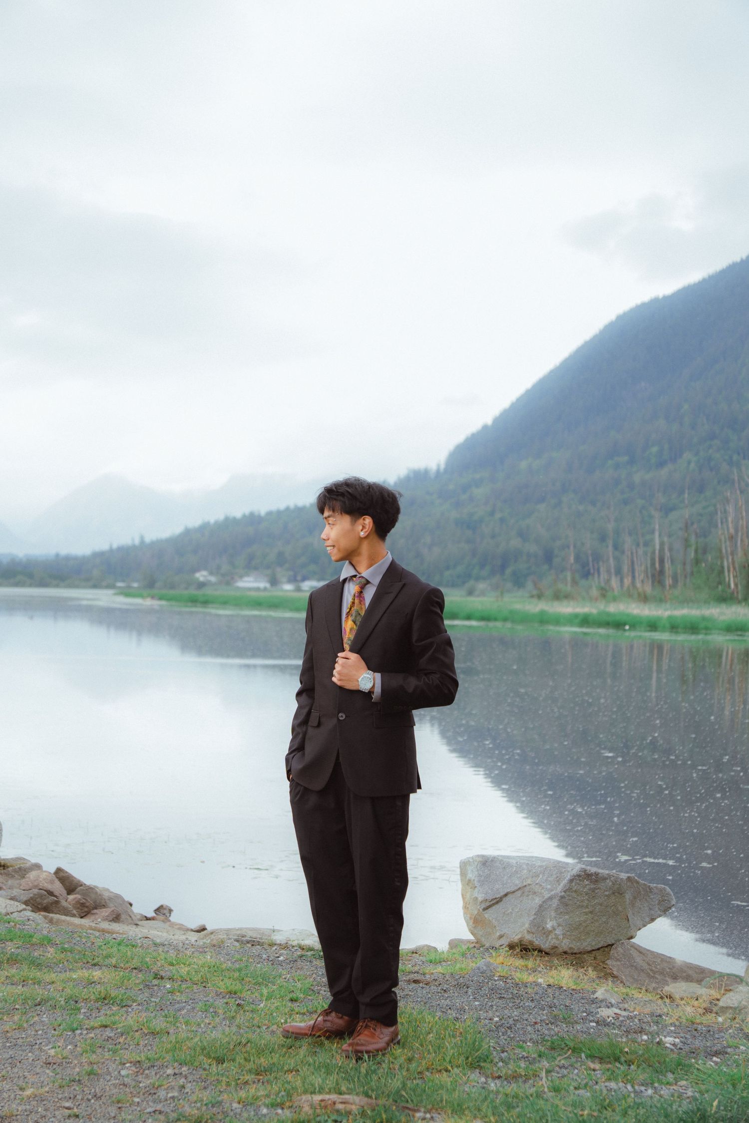 A young man in a dark suit gazes thoughtfully beside a serene mountain lake.