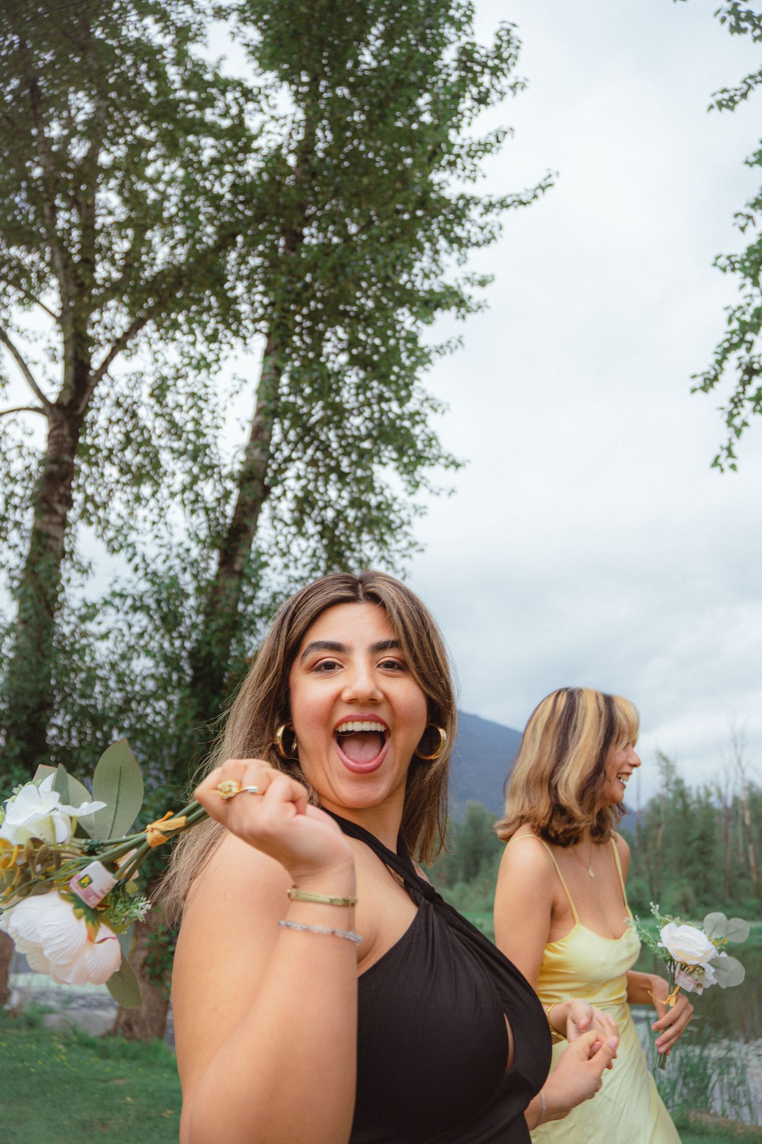 Excited young woman in black dress holding white flowers laughing outdoors by a lake with mountains.