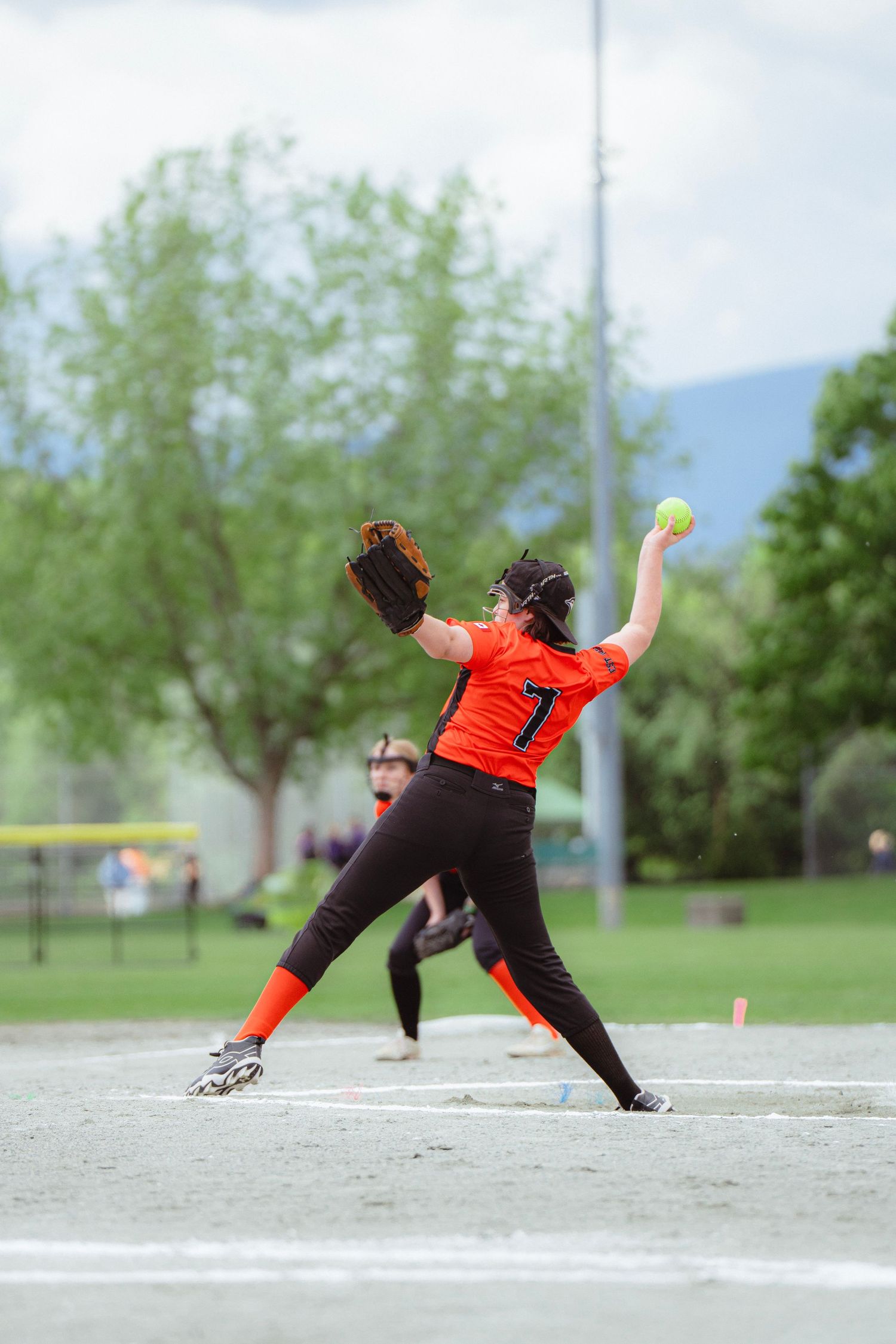 Softball pitcher wearing number 7 in orange uniform throws a yellow ball during an outdoor game.