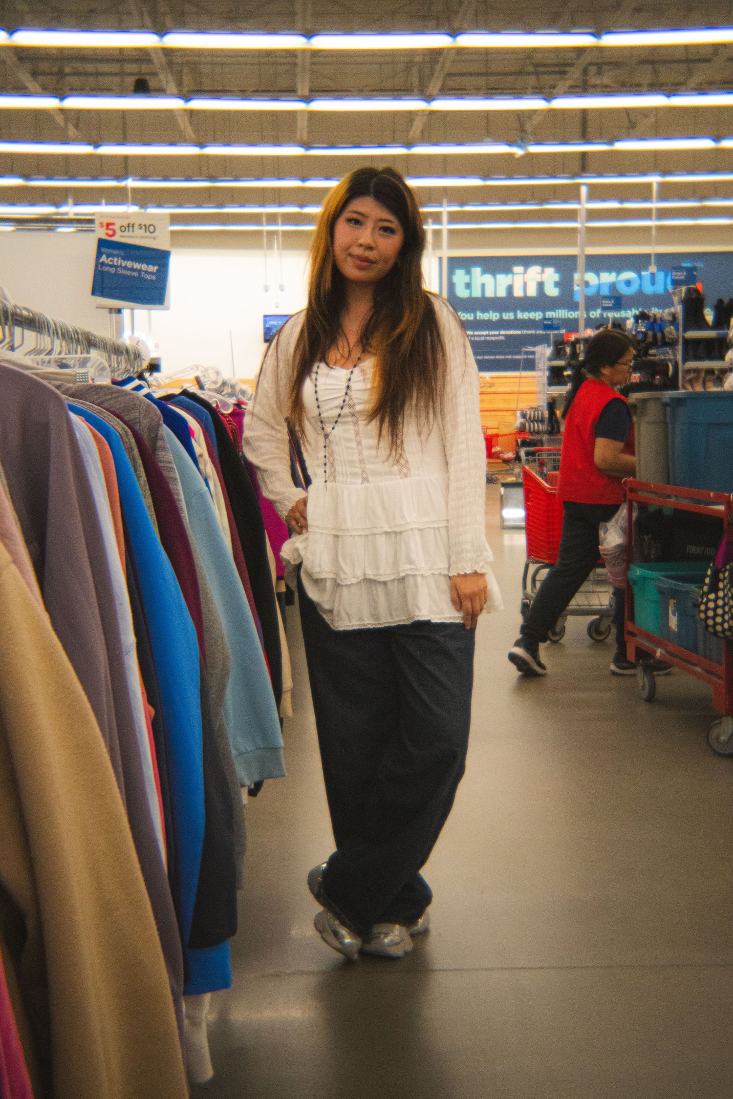 Woman in white ruffled blouse and dark wide-leg pants shopping in a brightly lit thrift store.