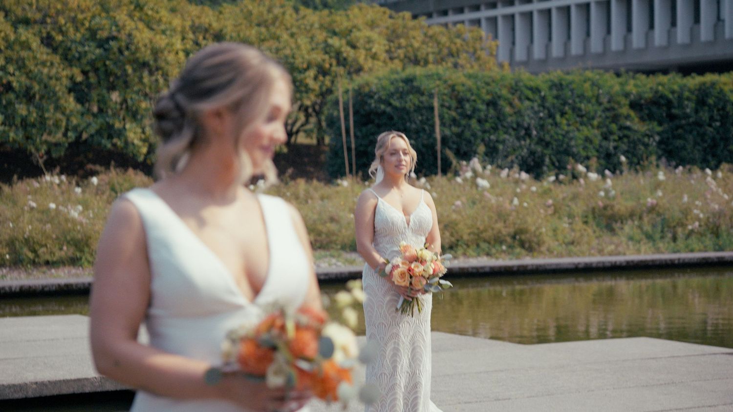 Two brides in white gowns holding bouquets outdoors near a reflecting pool.