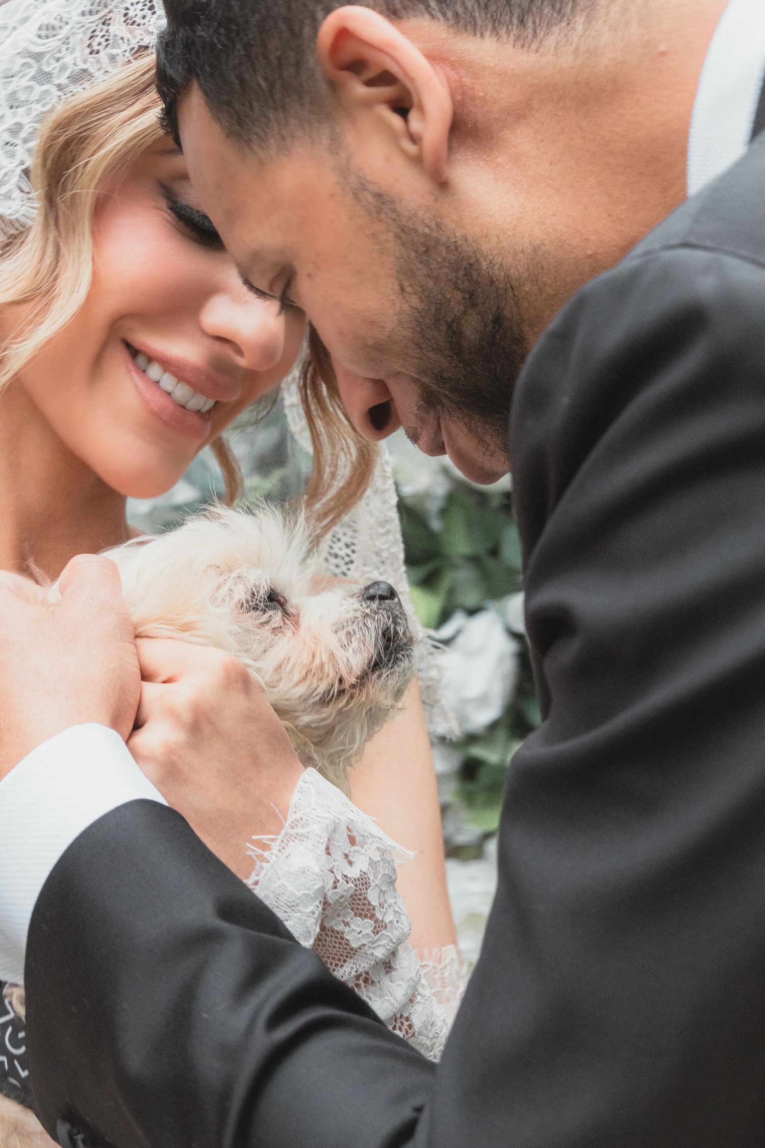 Bride and groom in wedding attire smile together while holding a small fluffy dog between them.