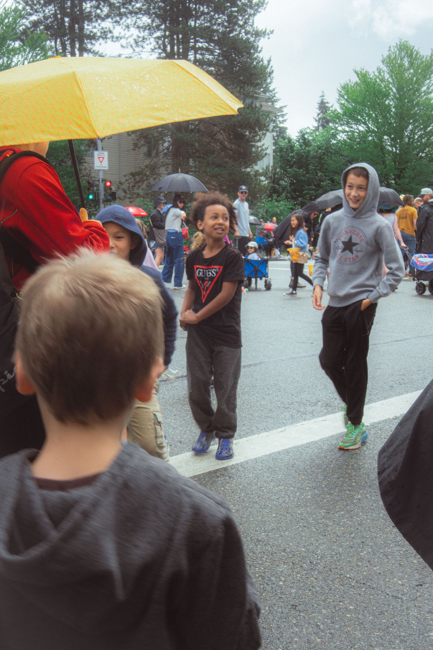 Children walk and watch a street parade under yellow umbrellas on a rainy overcast day.