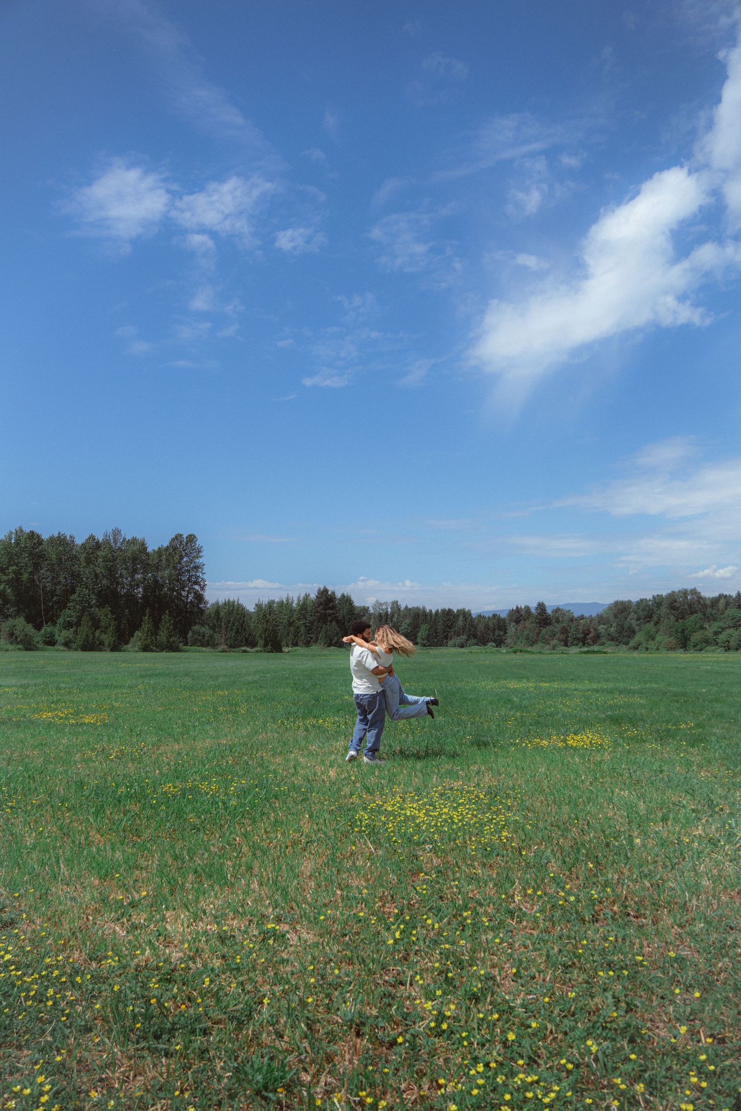An interracial couple embracing and spinning joyfully in a vast green meadow with wildflowers under a bright blue sky.
