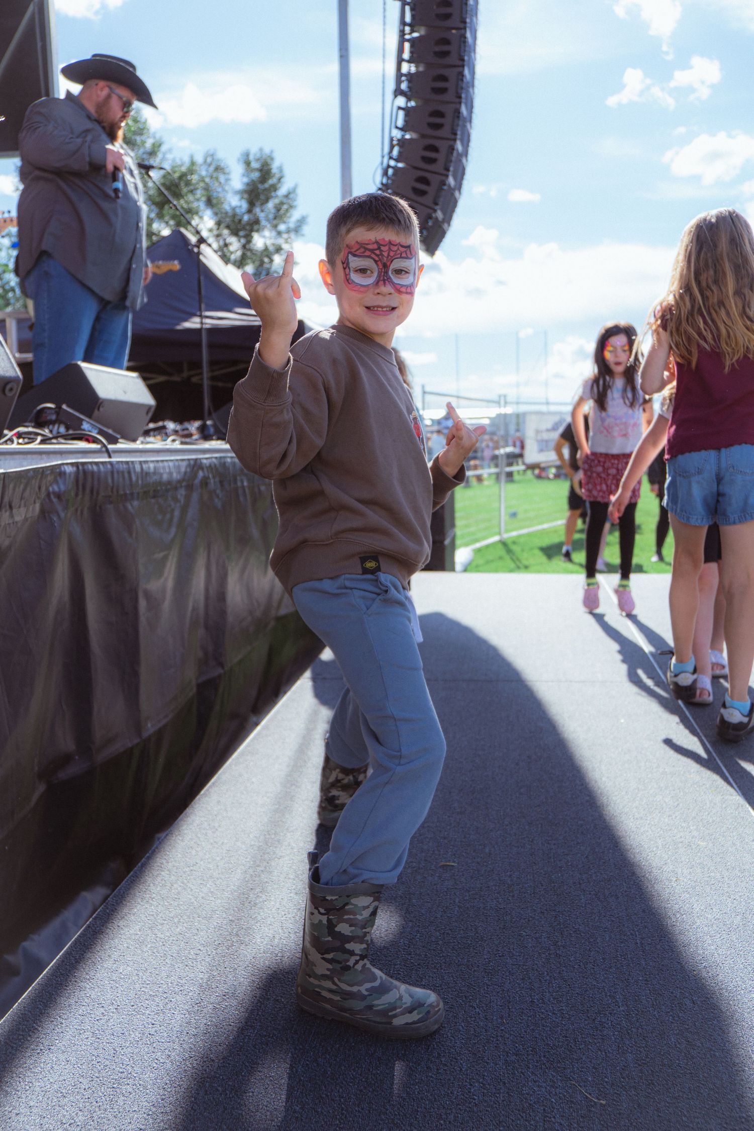 Young boy with face paint dancing near outdoor festival stage wearing camo boots and brown sweater.