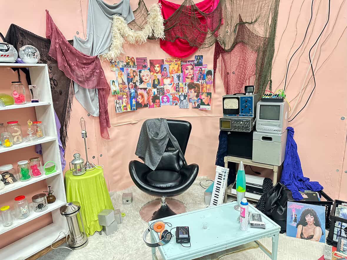 Colorful art installation room with retro TV, keyboard, shelves of bottles, and pop art wall collage.