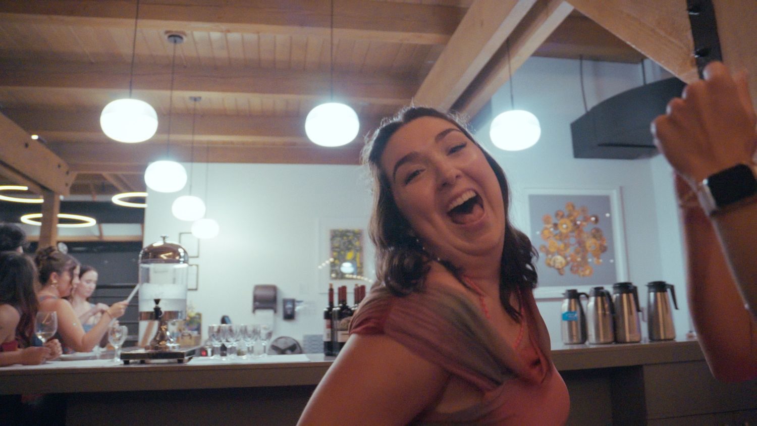 A laughing woman in a red top holds a camera in a bright modern cafe with pendant lights.