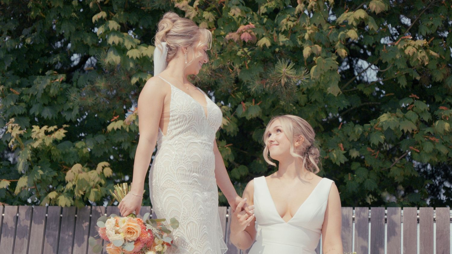 Two brides in white wedding dresses hold hands smiling outdoors with trees behind them.
