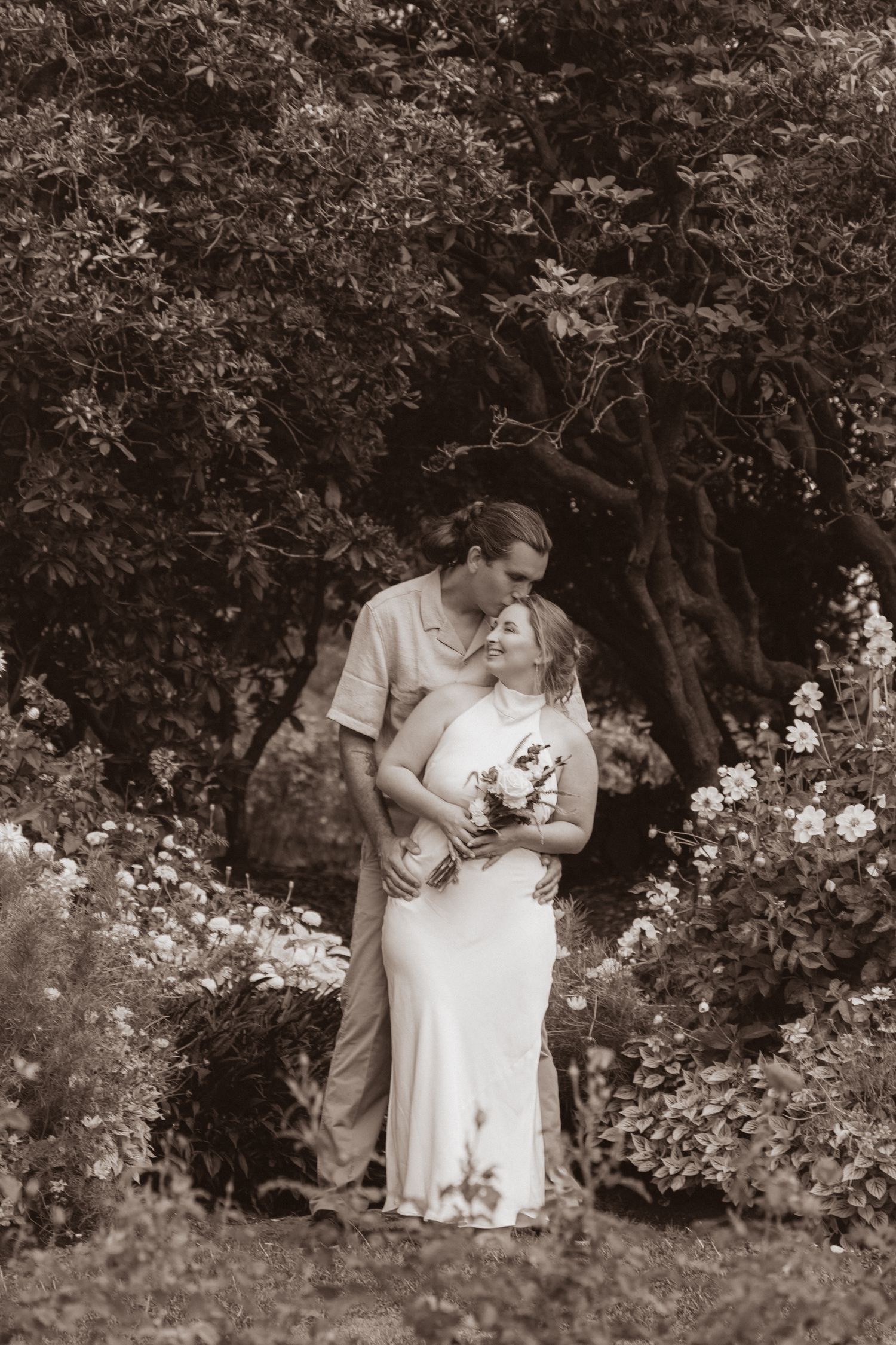 Romantic sepia-toned wedding photo of a couple embracing in a lush garden, groom kissing bride holding a bouquet.