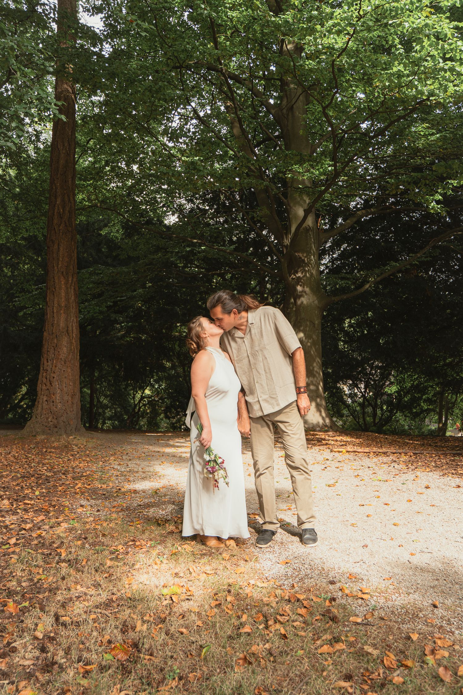 Newlyweds sharing a tender kiss on a scenic woodland path with towering trees.