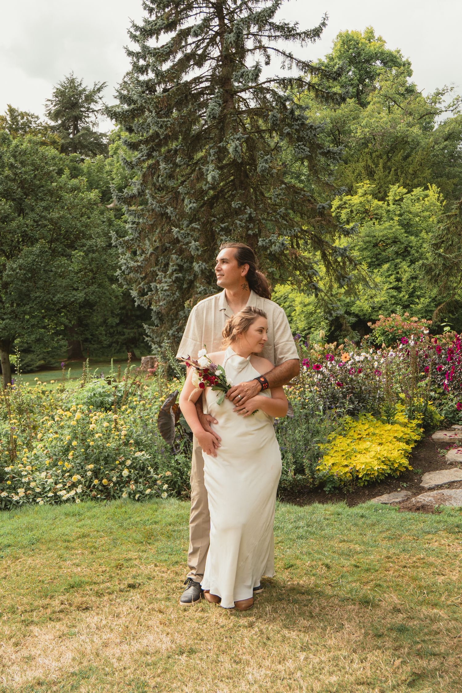 Groom holds bride from behind as they stand in a lush garden, bride holding a floral bouquet in wedding attire.
