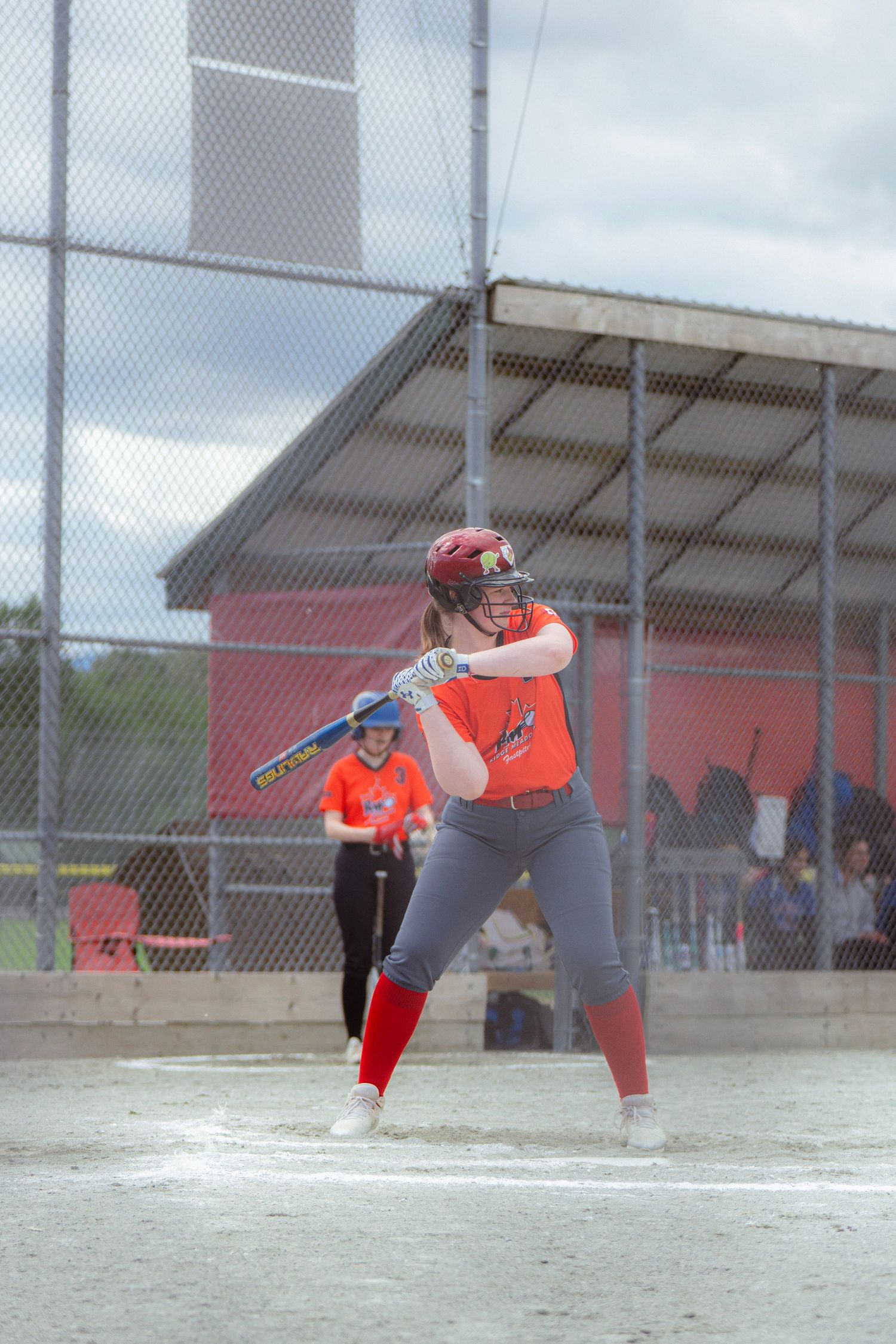 Female softball player in orange jersey and red socks assumes batting stance at home plate.
