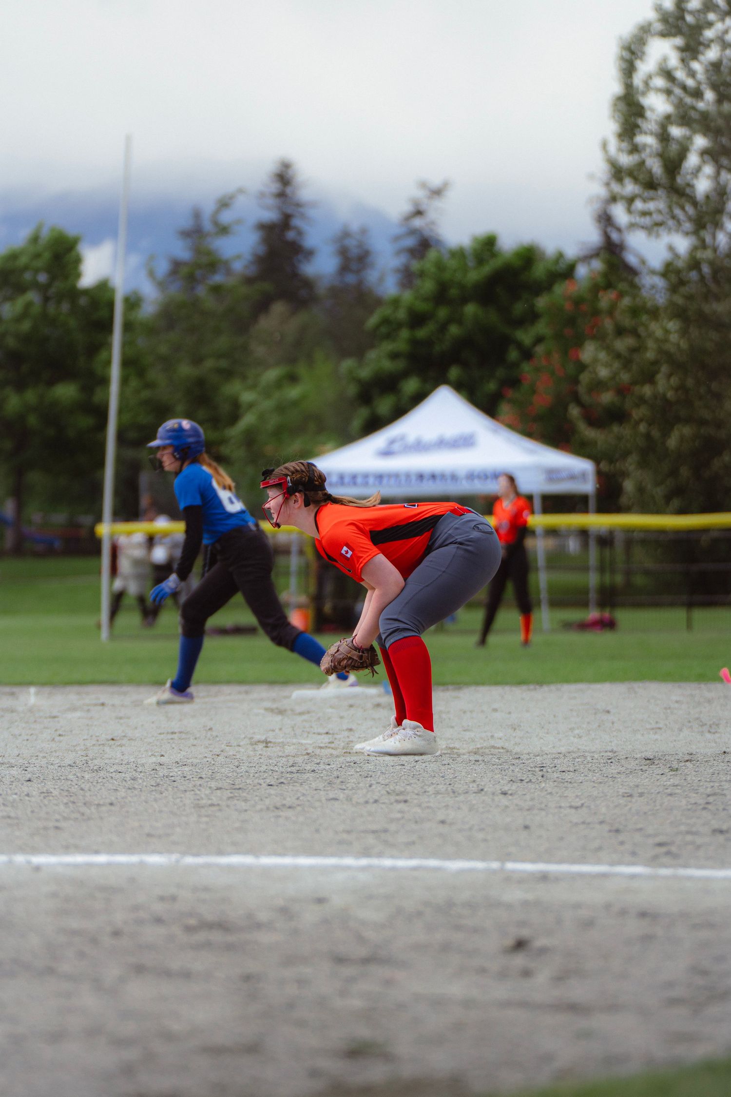 Softball fielder in orange uniform bends to field a ground ball during a youth game on a cloudy day.