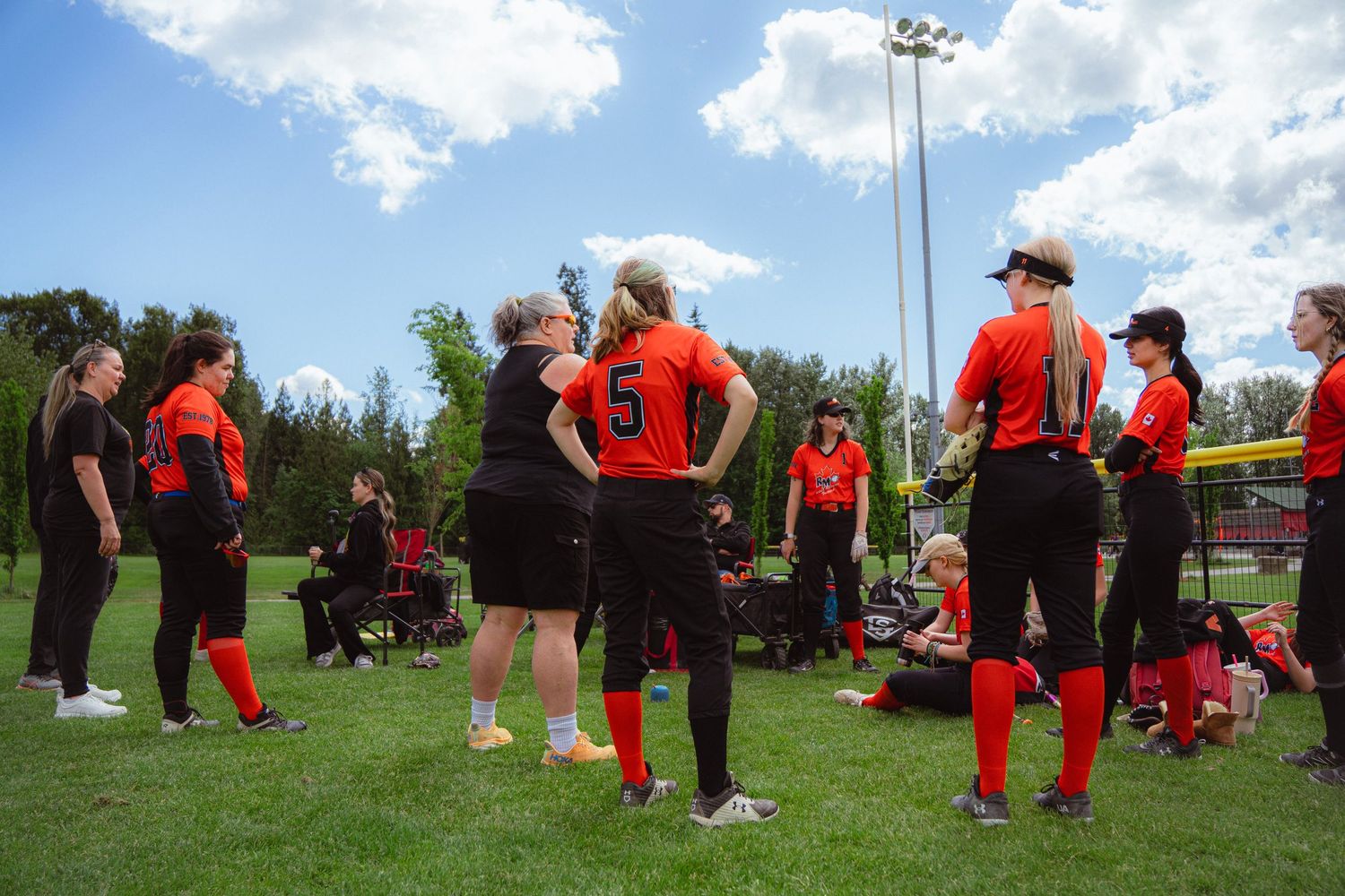 Female softball team in orange and black uniforms gathered on green field during sunny day.