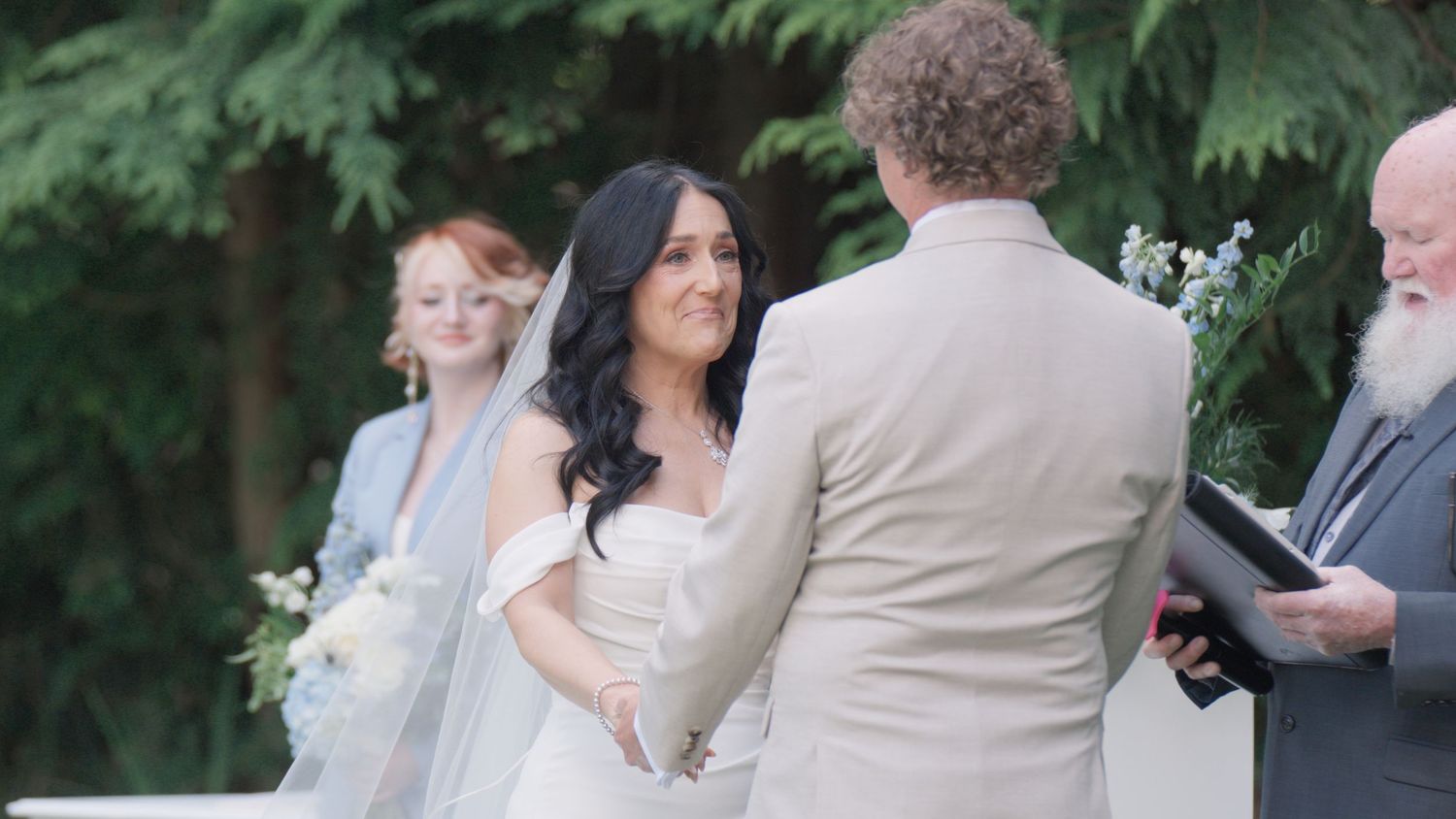 Bride in white off-shoulder gown holds hands with groom in tan suit during outdoor wedding ceremony.