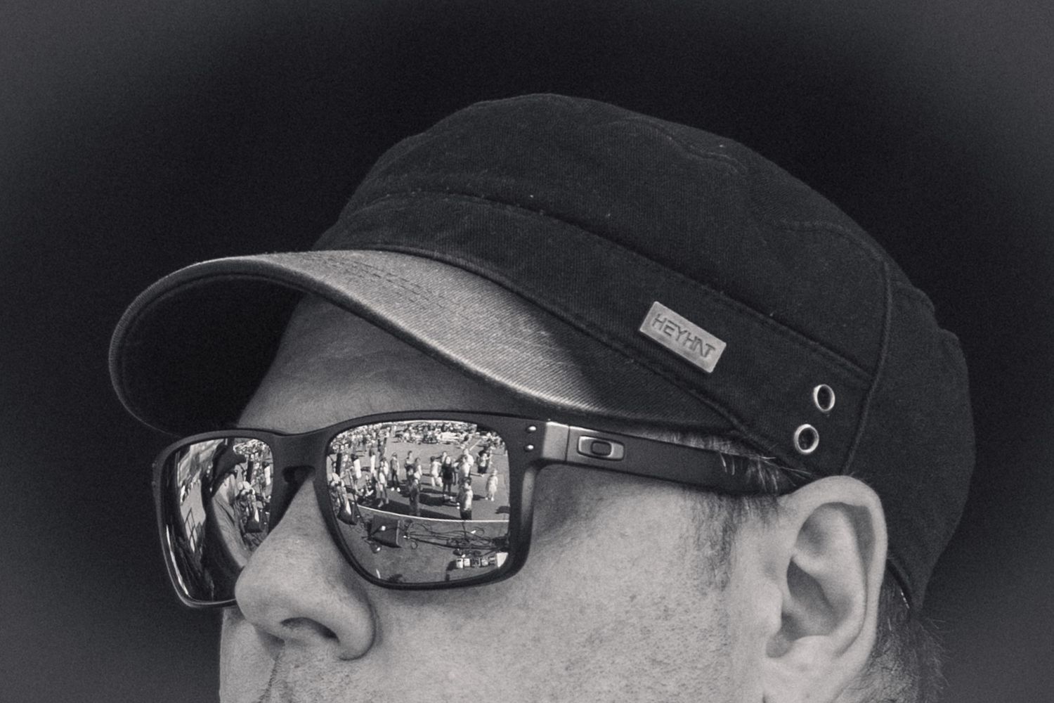 Black and white close-up of man in cap and sunglasses reflecting crowd at outdoor music event.
