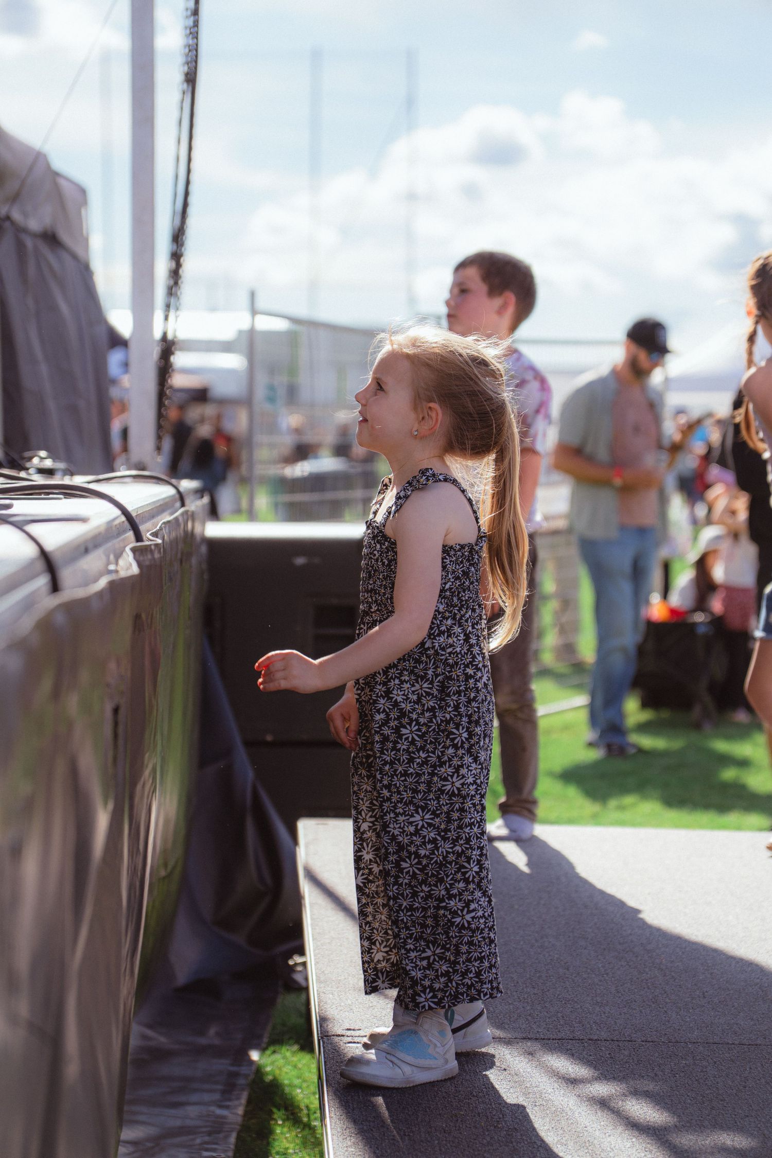Young girl in floral dress standing near outdoor concert stage watching performance.