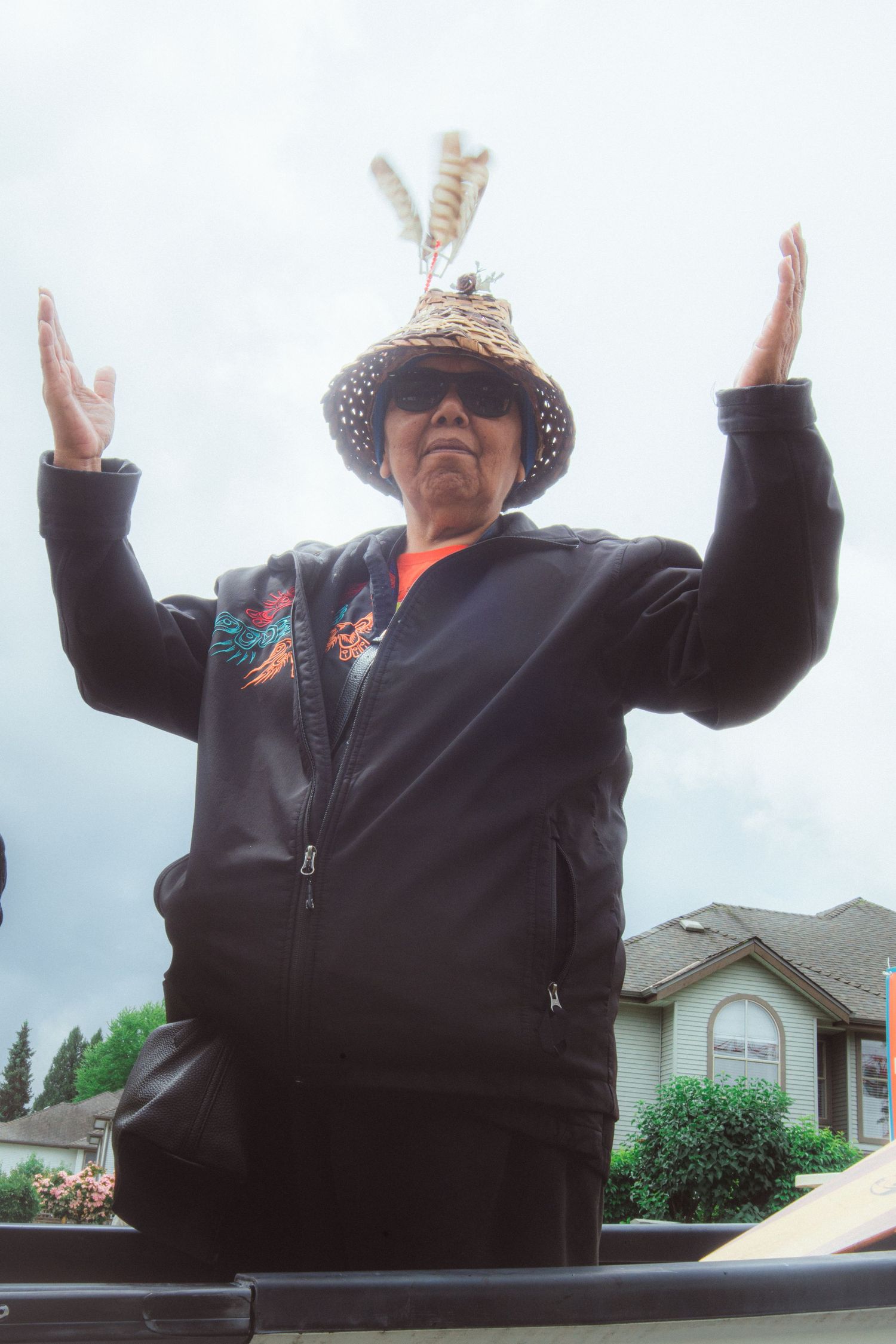 Indigenous elder in woven hat raises both hands skyward while standing on a parade float.