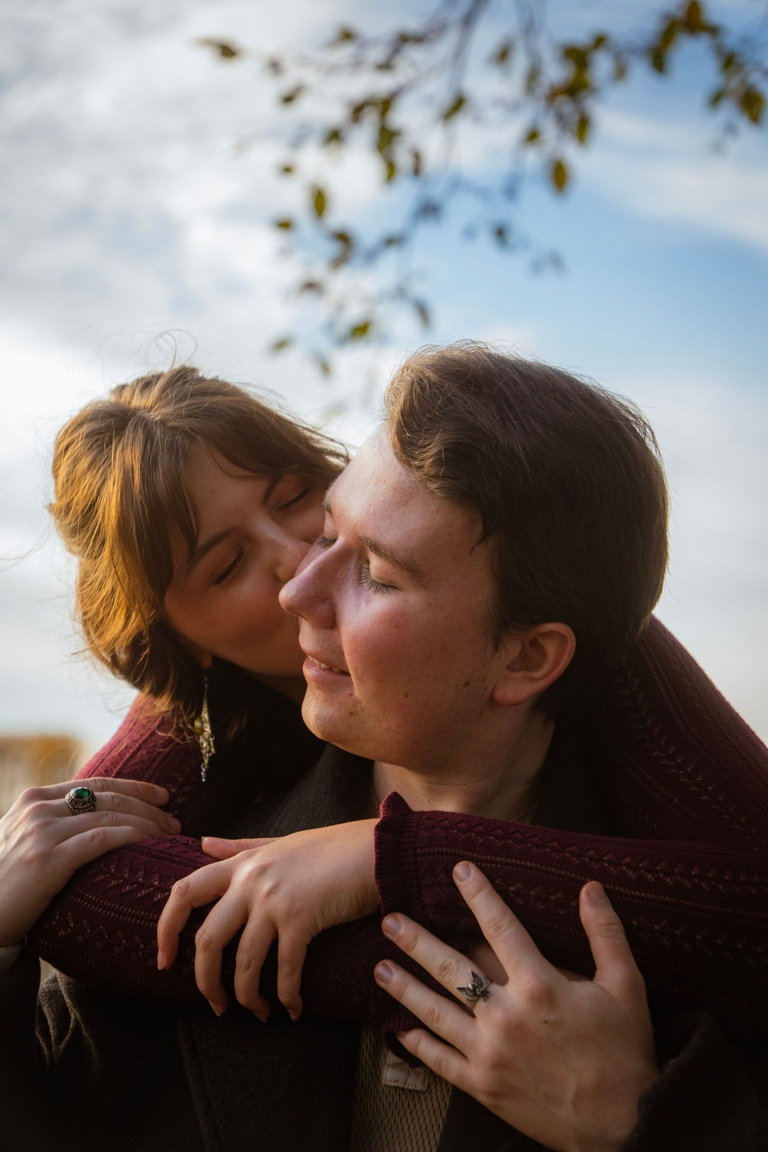 Woman tenderly kissing mans cheek while embracing him from behind under autumn branches.