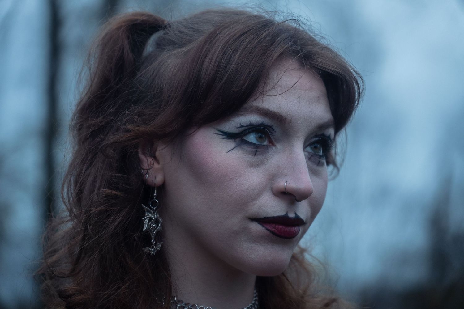 A woman with dramatic gothic makeup, dark lip color, and ornate earrings poses outdoors in a moody twilight setting.