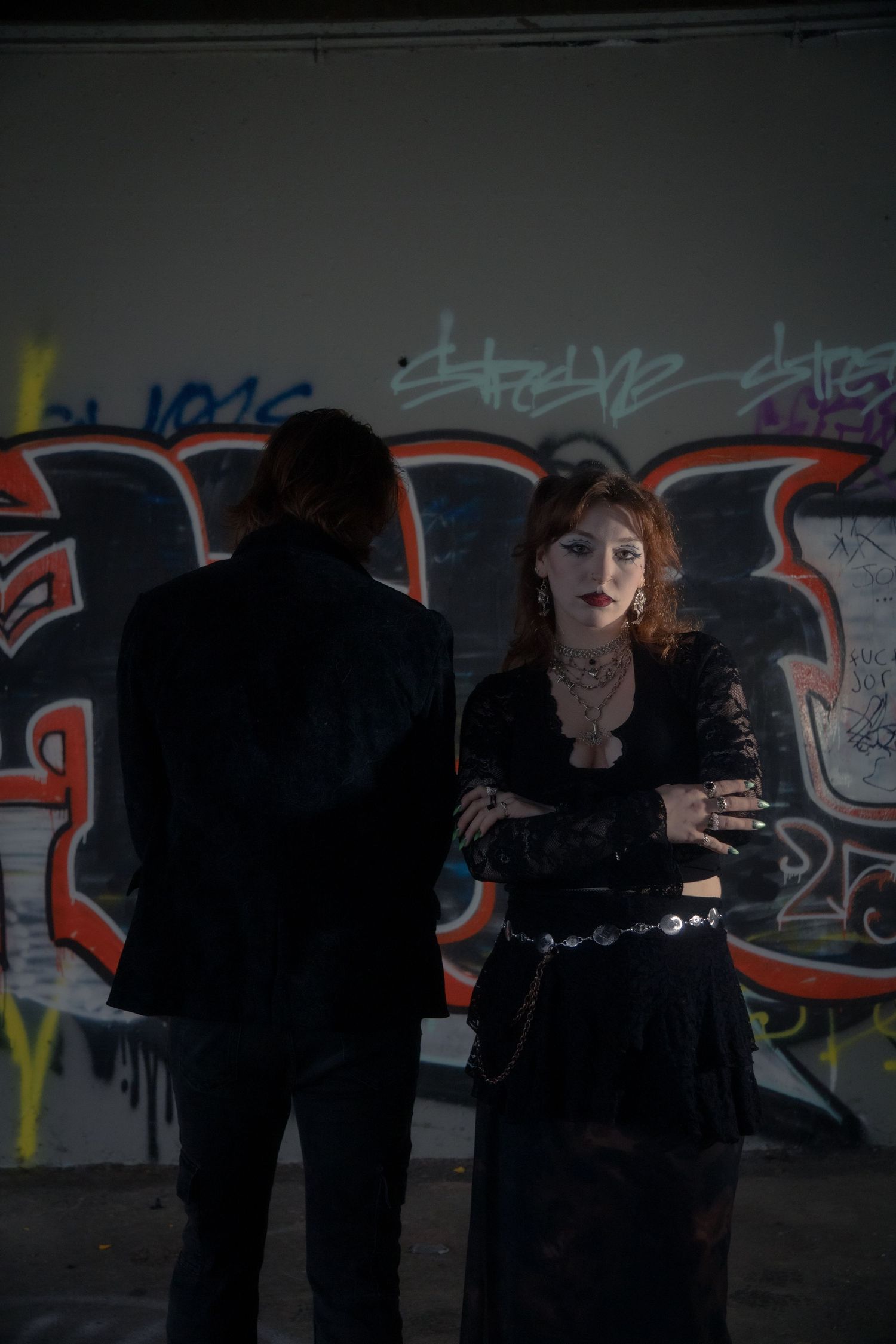 Gothic woman in black lace top with arms crossed stands before colorful graffiti wall.