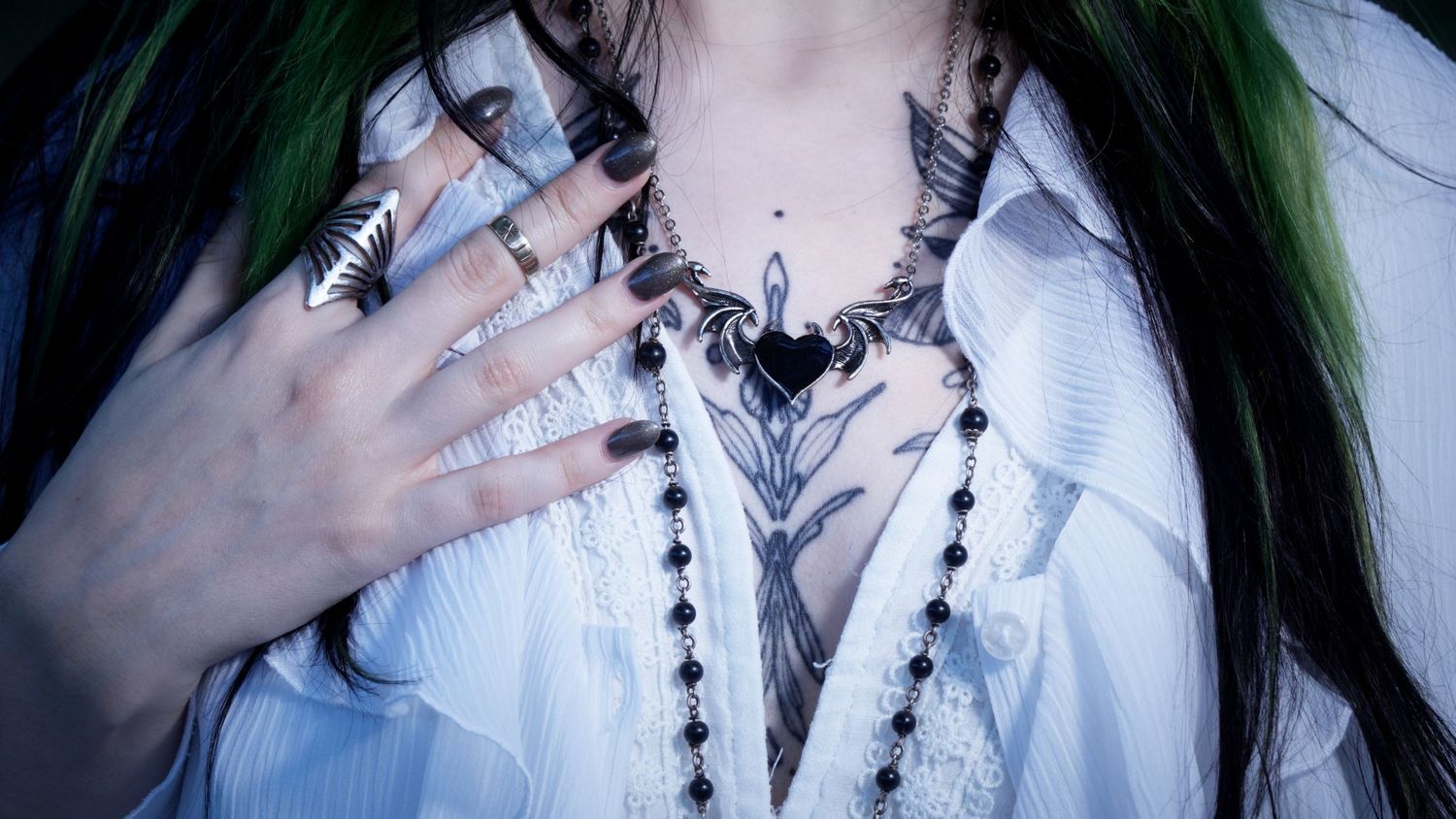 Close-up of chest tattoo with heart motif, black necklace, and ringed hand on white blouse.
