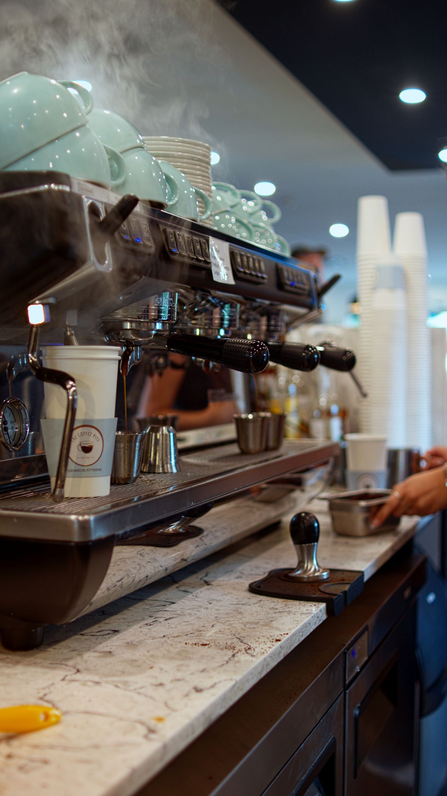 Busy cafe espresso bar with steaming professional coffee machine, stacked cups, and marble countertop.