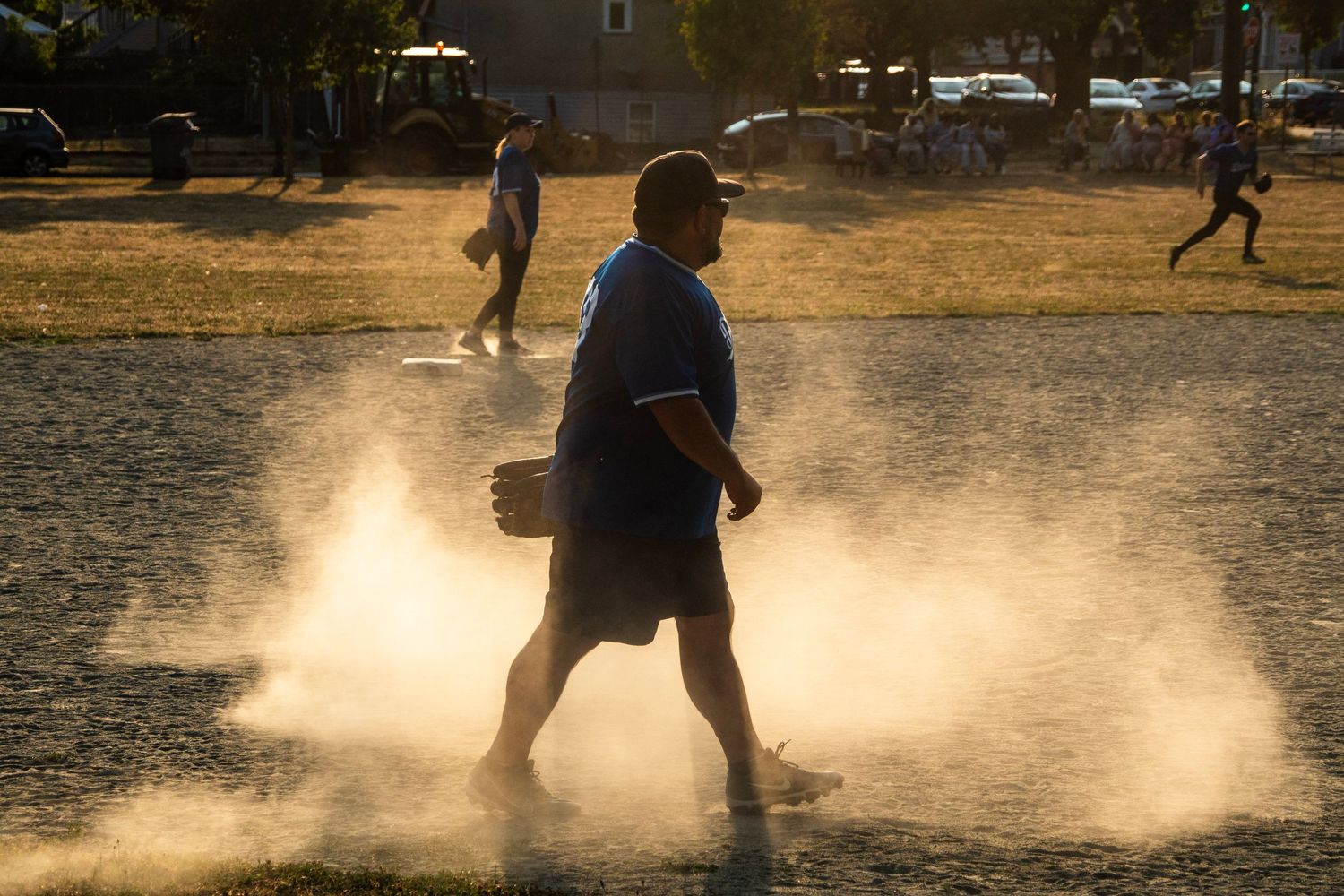 Man walking through a dusty softball field at sunset with players visible in the golden light background.