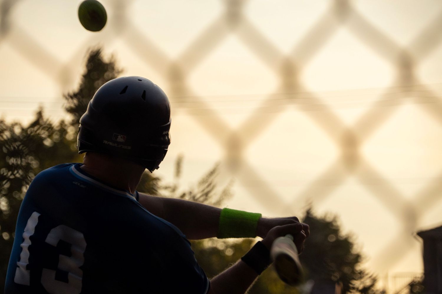 Silhouetted baseball batter in helmet swings at a pitch viewed through a chain-link fence at dusk.
