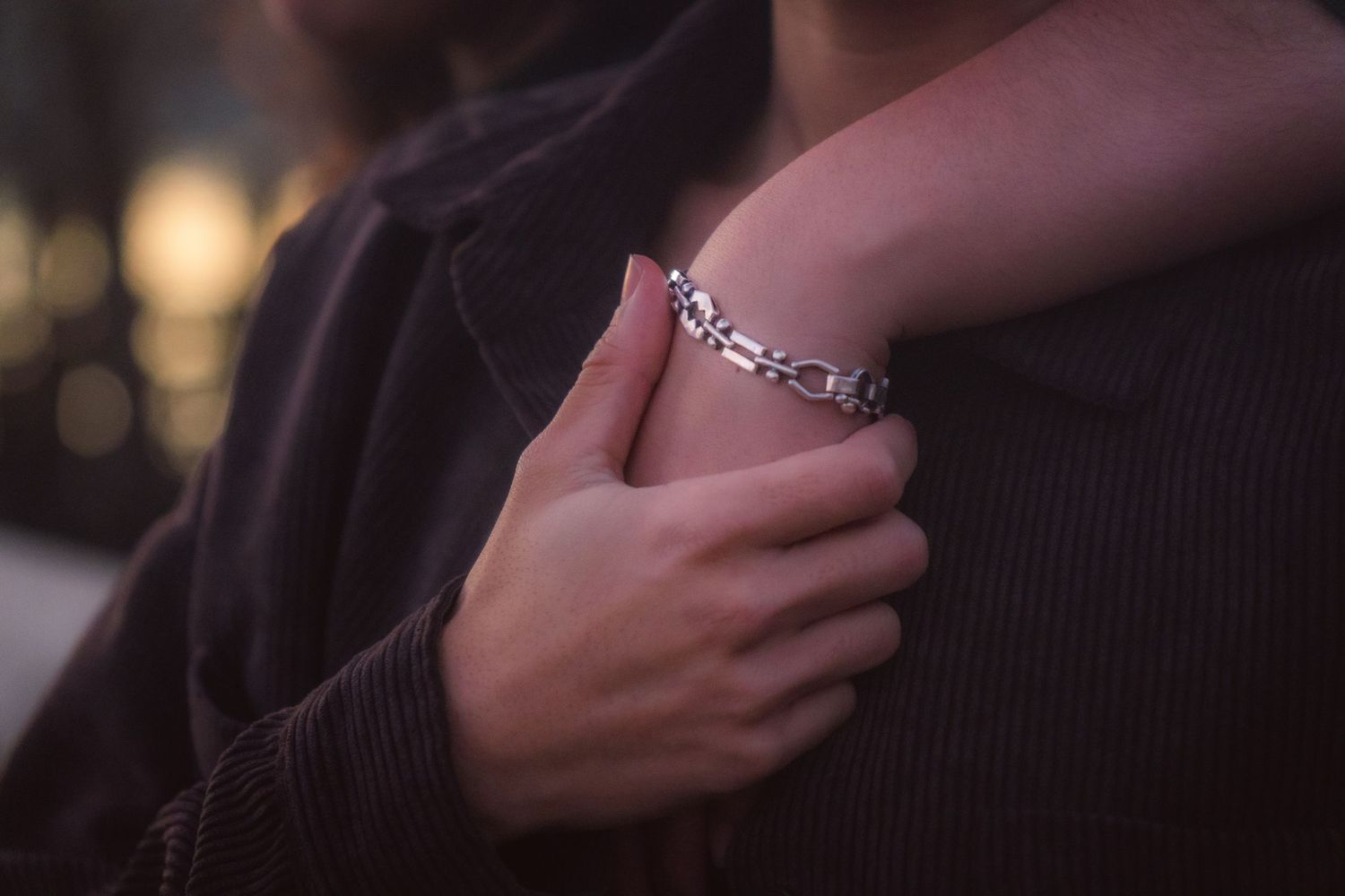 A close-up of a hand with a silver chain bracelet gently resting on a partners brown jacket at dusk.