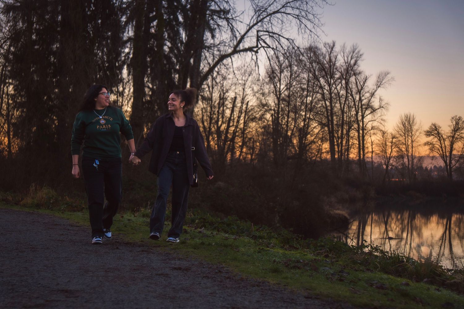 Two women walking hand in hand along a lakeside path at dusk.