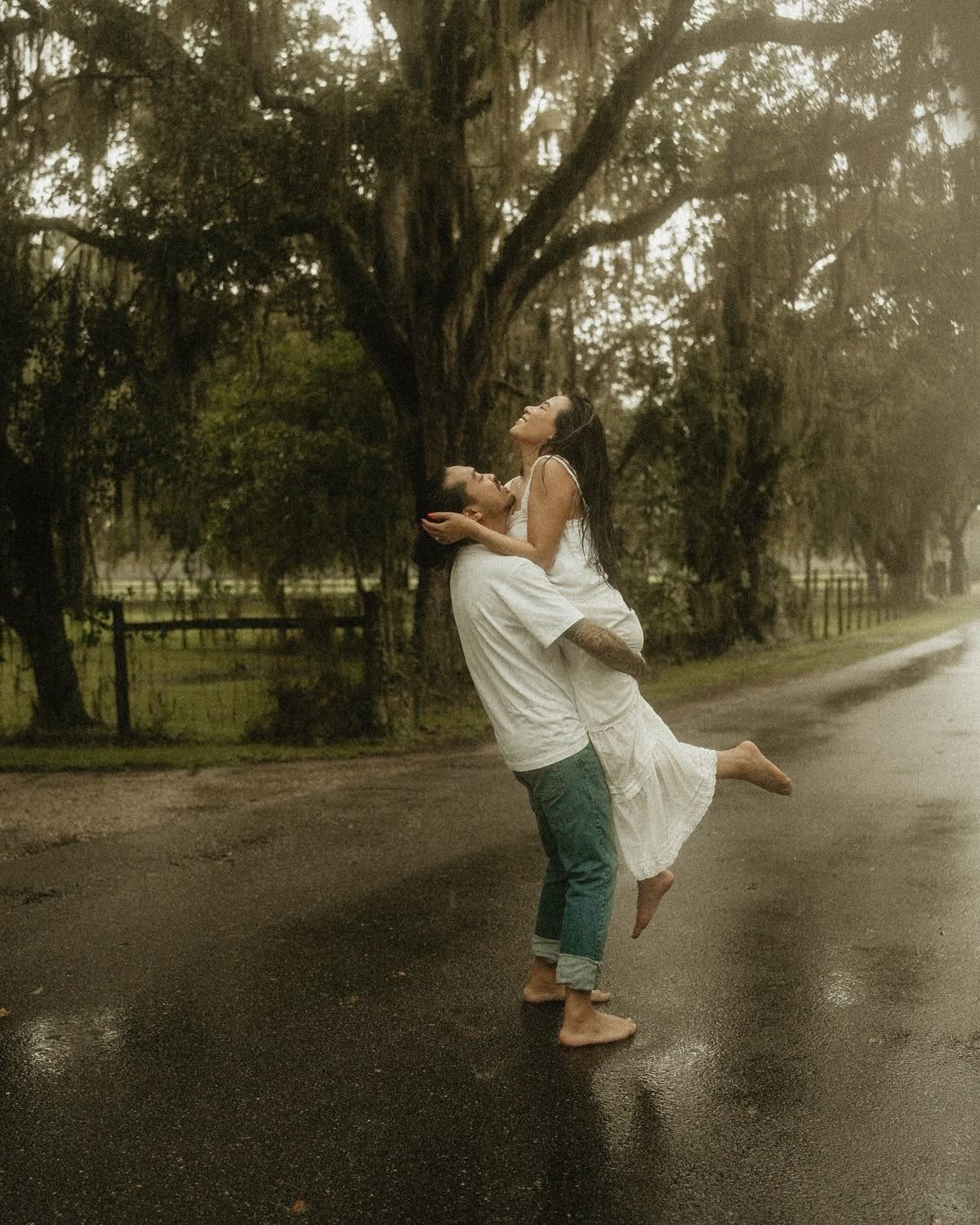 Couple dancing in the rain on a wet road surrounded by moss-draped trees, man lifting woman in white dress.