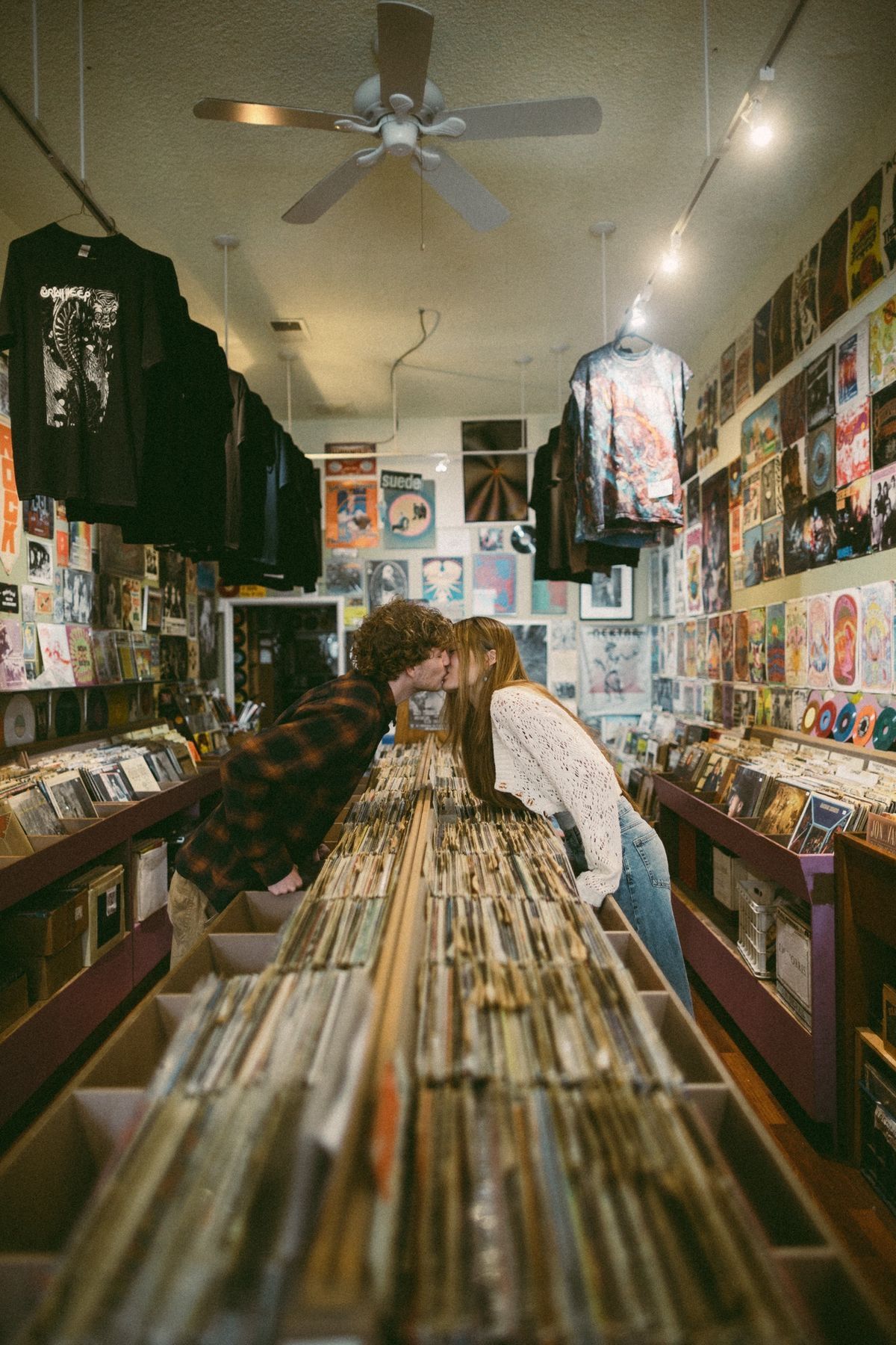 Couple sharing a kiss over vinyl record bins in a cozy, poster-filled record shop with ceiling fan above.