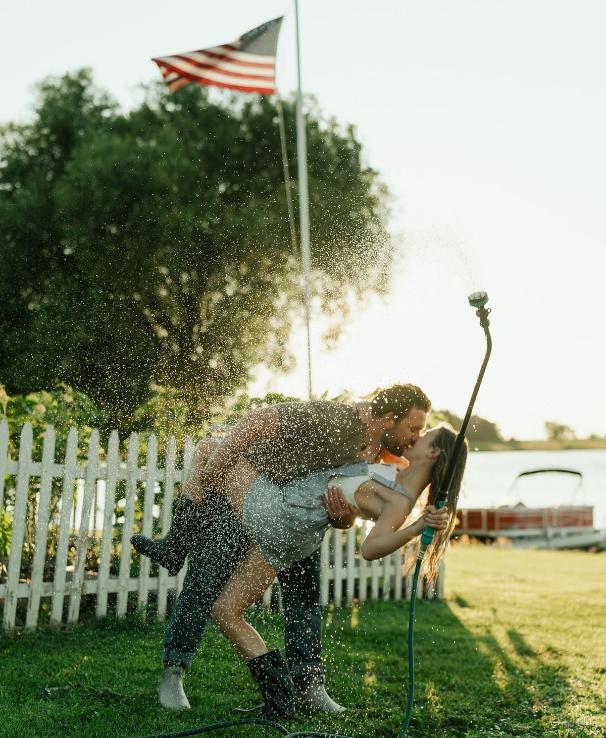 Couple kissing in sprinkler spray in backyard with American flag and white picket fence on sunny day.