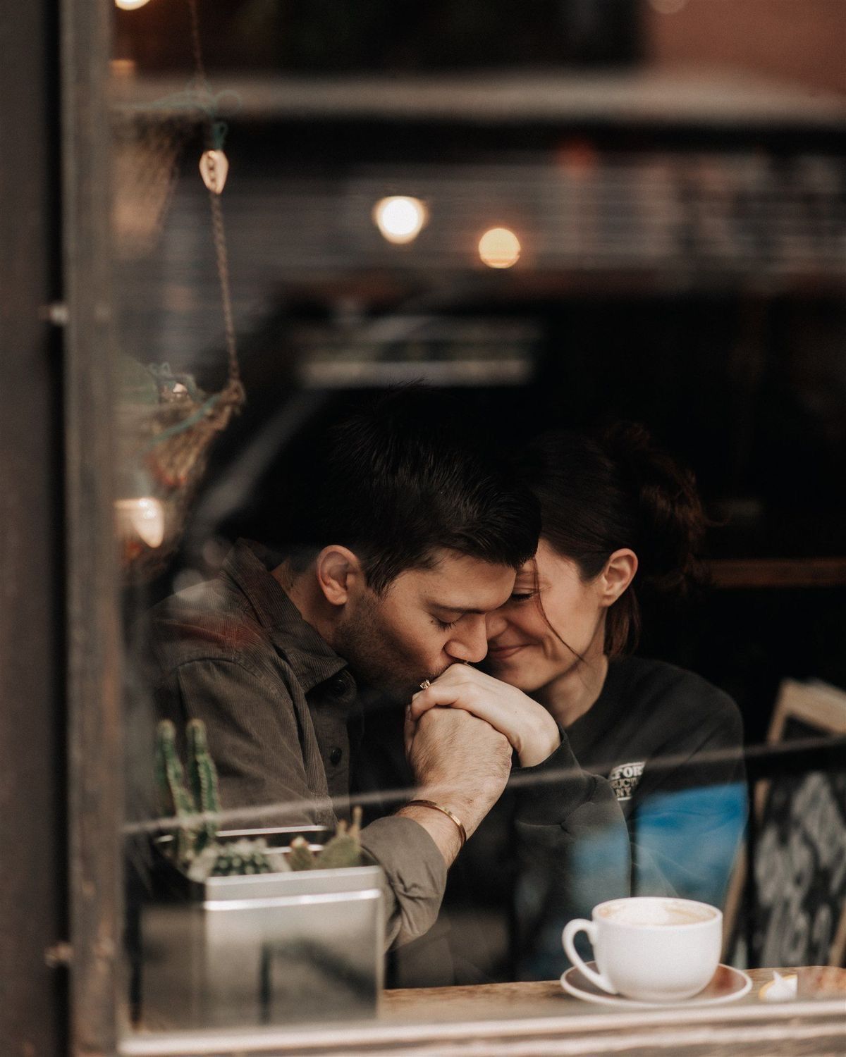 A loving couple holds hands and touches foreheads intimately at a cozy cafe window seat with coffee.