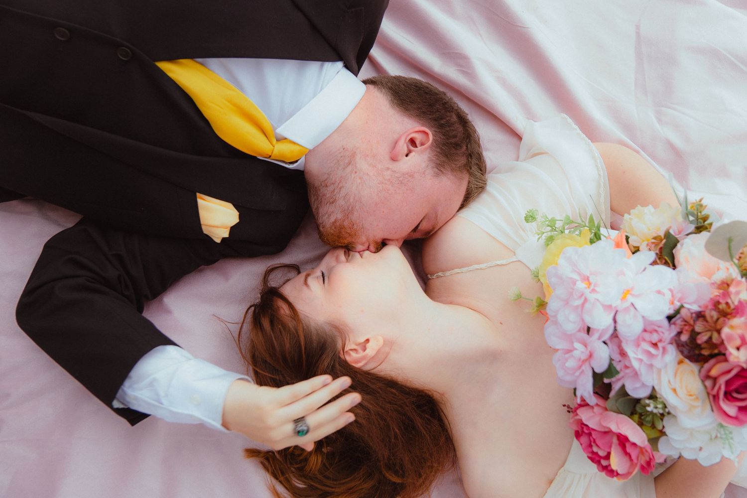 Newlywed couple lying on pink fabric, groom in black suit with yellow tie kissing bride on cheek.