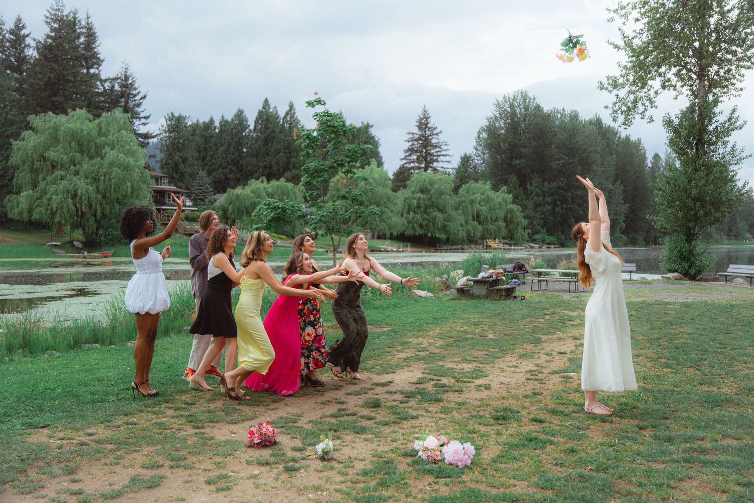Bride in white gown tosses bouquet to excited bridesmaids on green lawn by scenic lake.