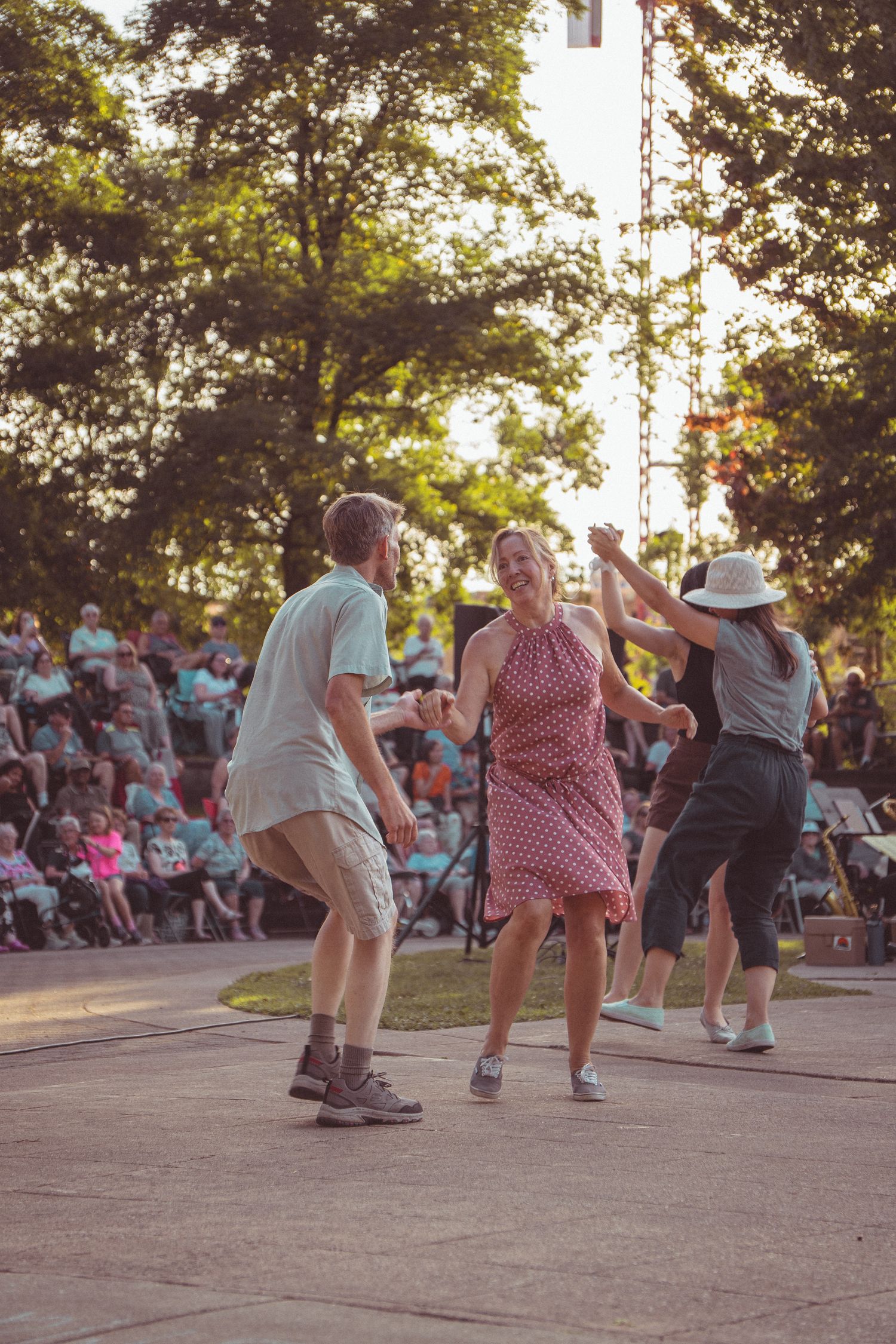 People dancing outdoors at a summer festival concert with an audience seated in the background among green trees.