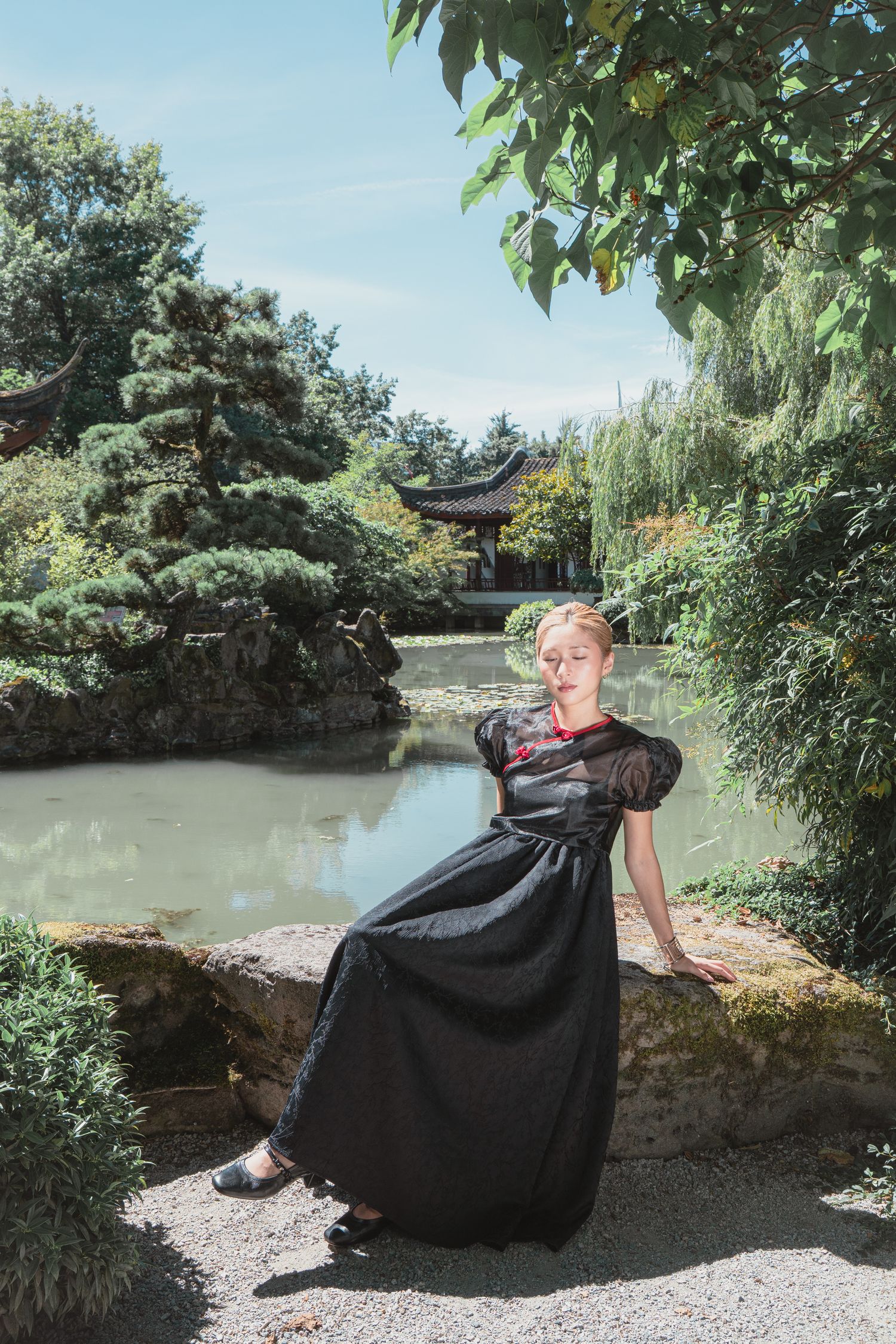 Woman in black dress poses by a tranquil pond in a Chinese garden with a pagoda and lush greenery.