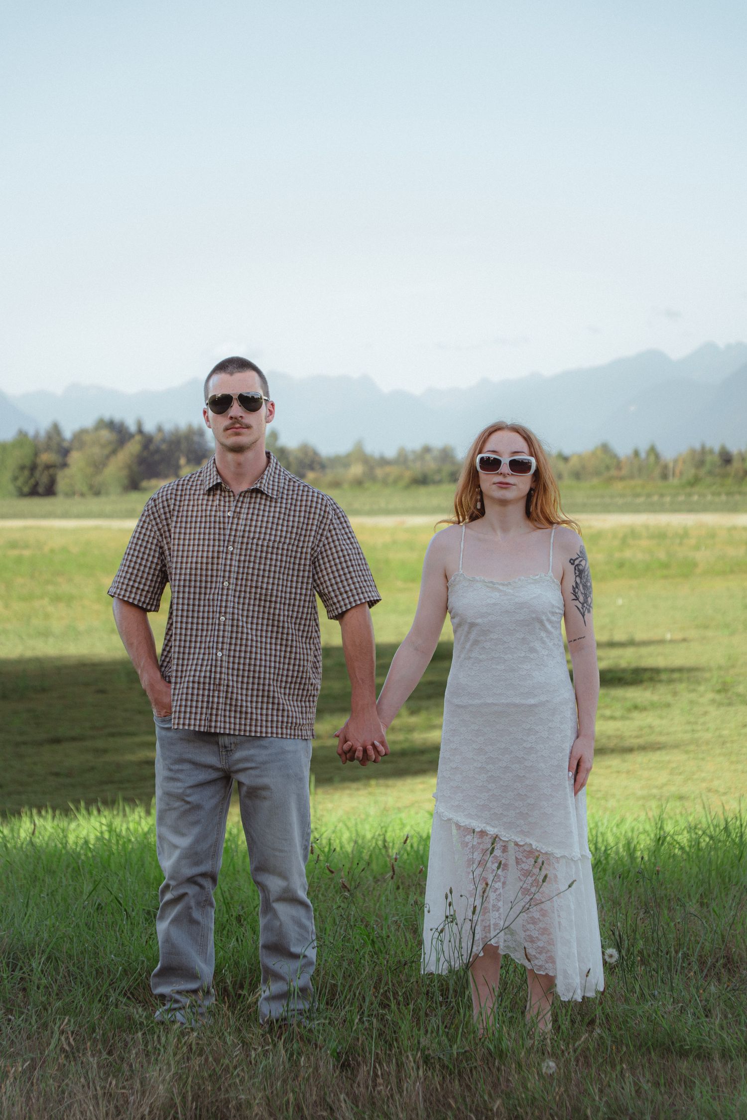 A couple holding hands and standing in a green meadow with mountain views, both wearing sunglasses.
