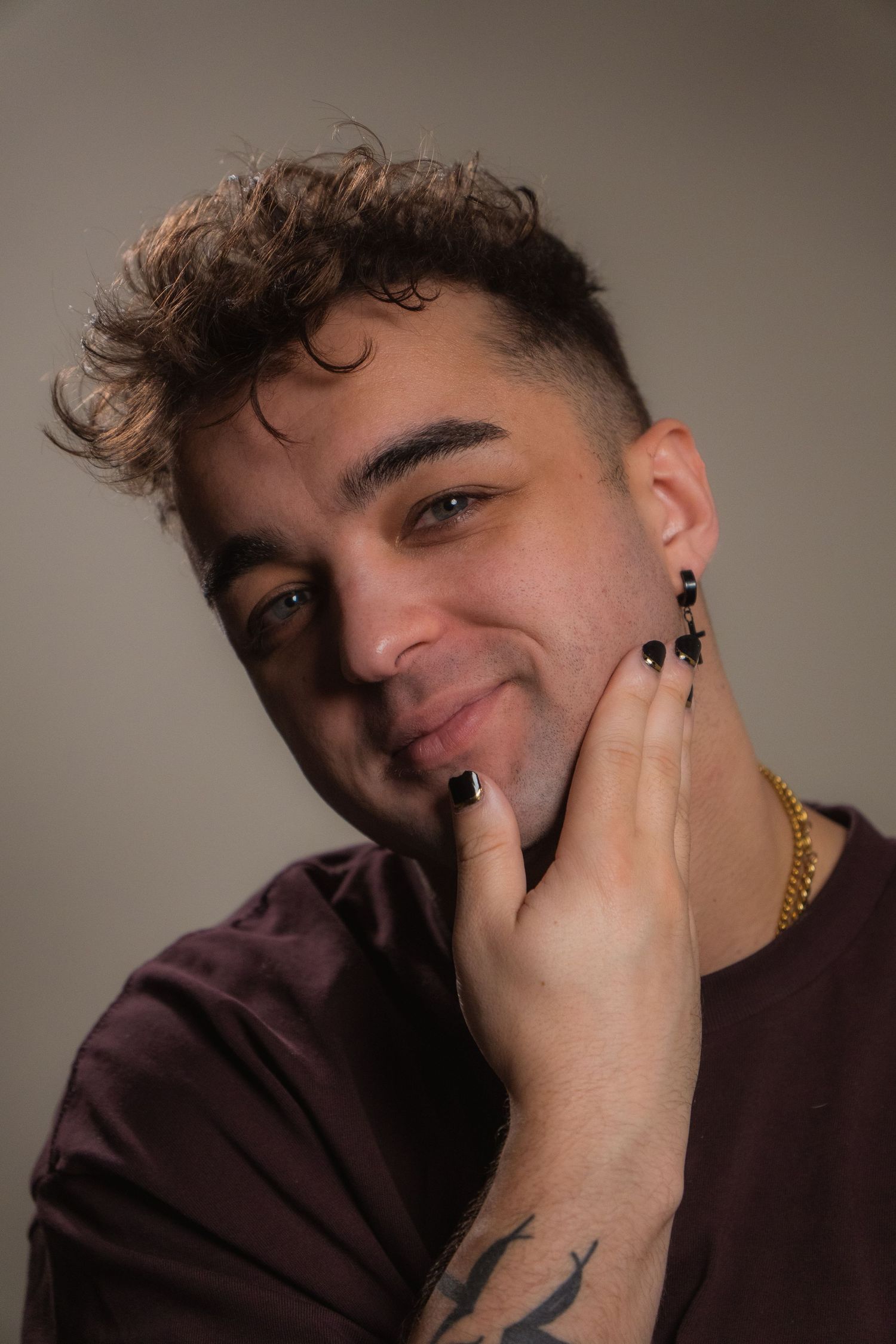 Smiling young man with curly hair and beard touching face, wearing gold chain and maroon shirt, tattoo visible.